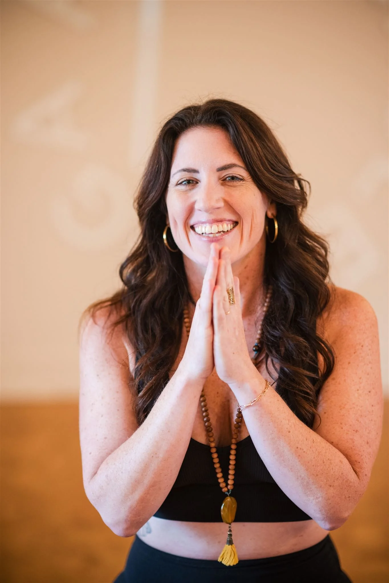 A woman with long, wavy brown hair, smiling and holding her hands together near her face. She is wearing hoop earrings, a beaded necklace, and a black sleeveless top. The background is neutral and blurred.