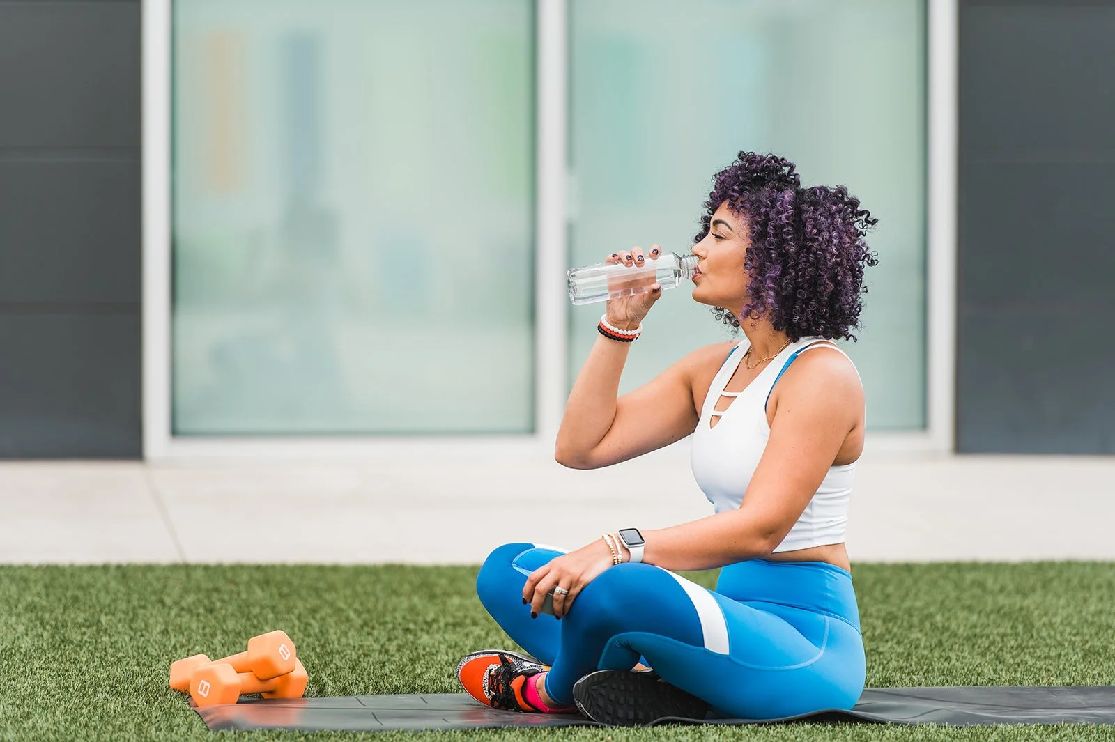 Woman with purple curly hair in workout attire sitting on yoga mat, drinking water during outdoor exercise session with dumbbells and fitness equipment nearby.