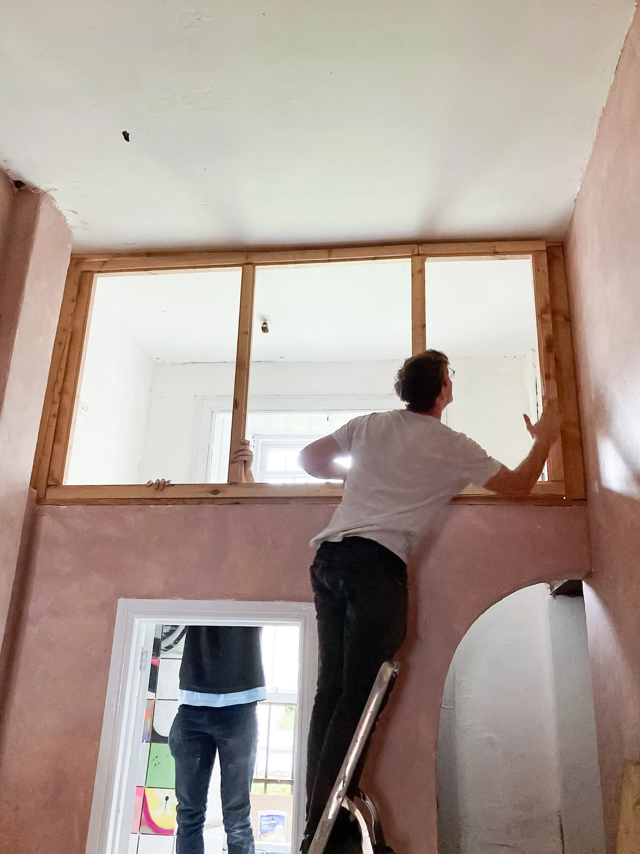 A person standing in a doorway, holding onto a wooden frame and gesturing with their hand. The room has a large window and visible walls with some artwork.