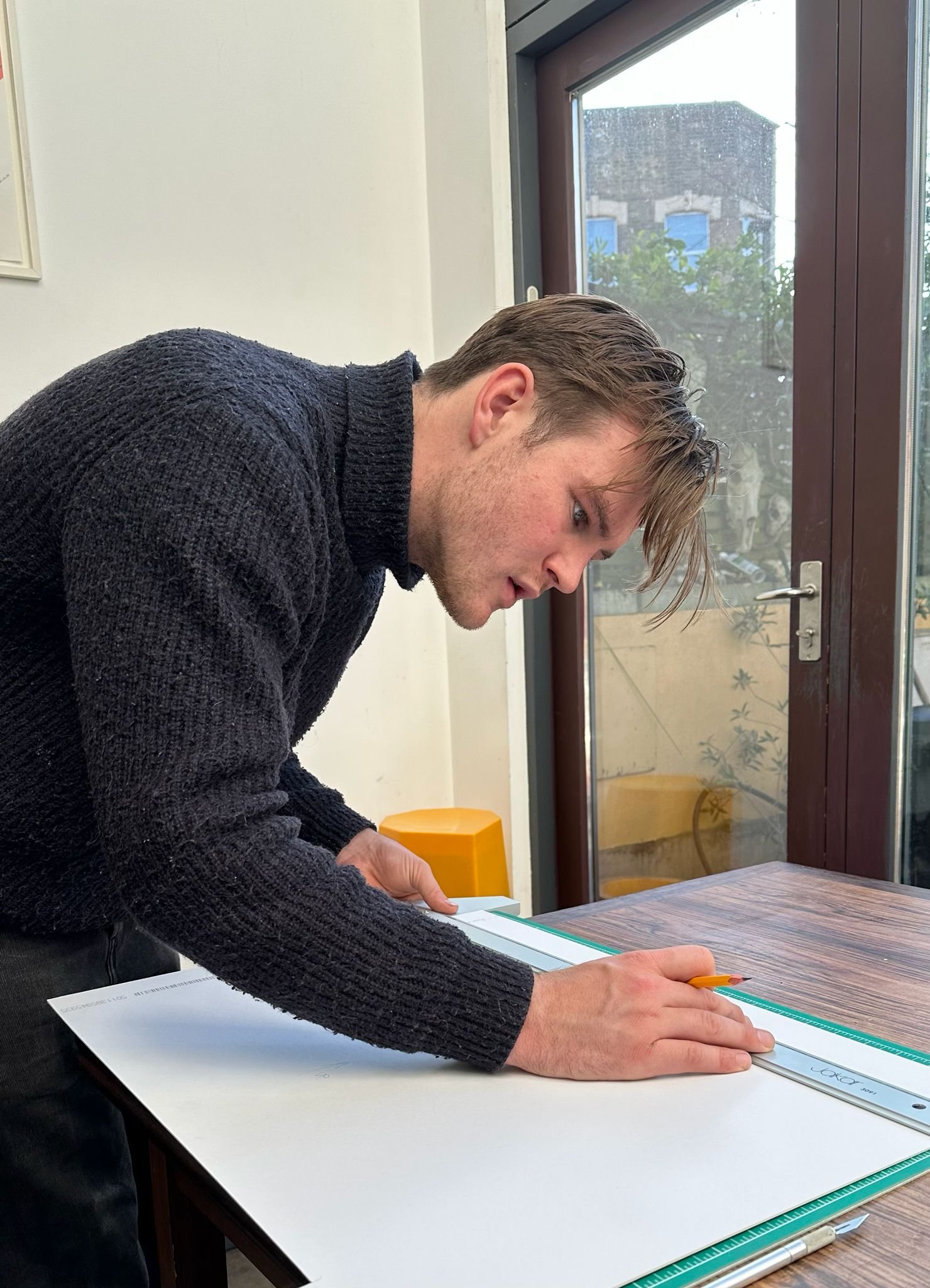 A man wearing a dark sweater is working on a drafting table, using a ruler and a pencil, in a room with large windows and a view outside.