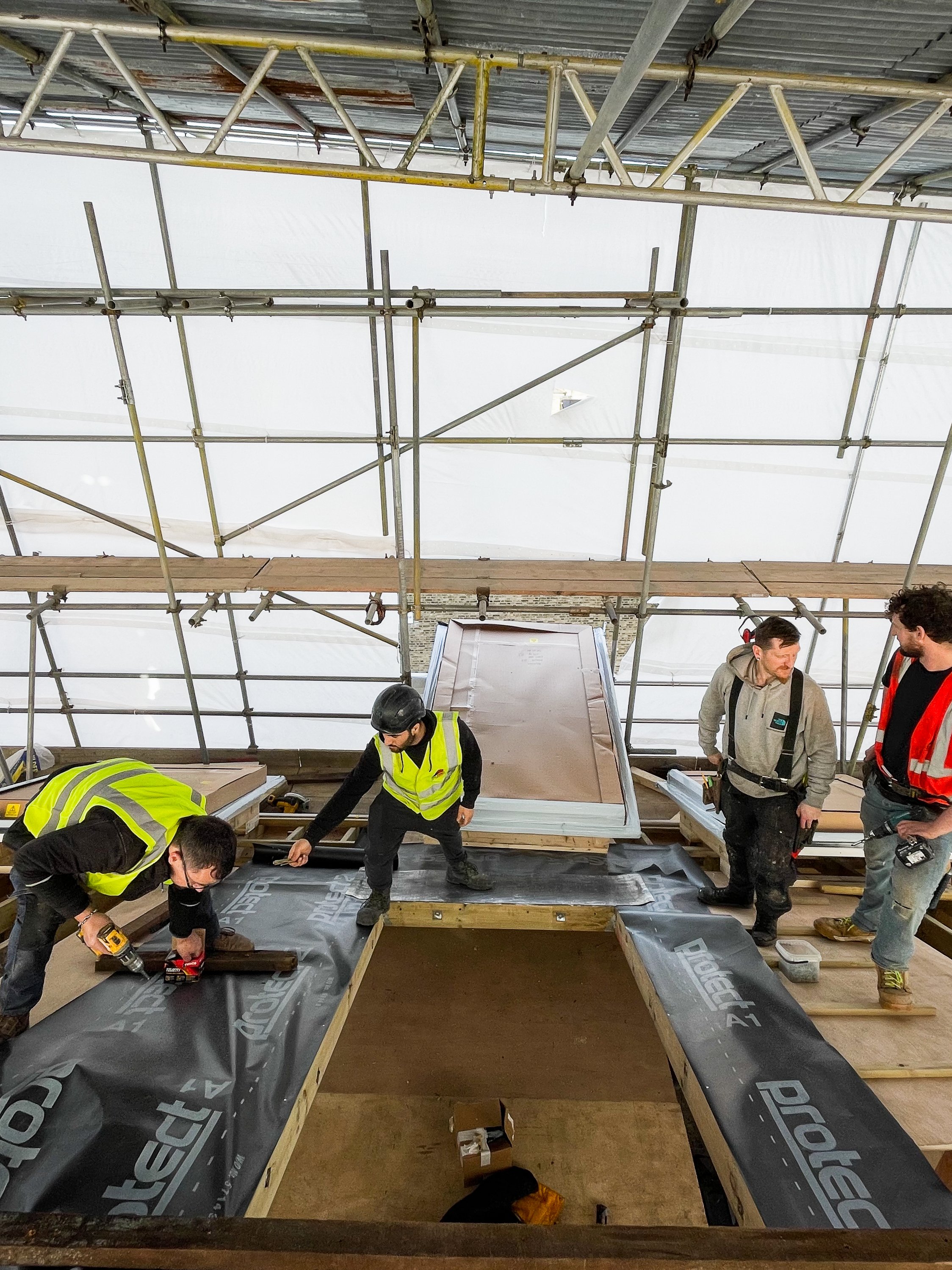 Construction workers install waterproofing material on the roof of a building under construction. The roof is surrounded by scaffolding, and the workers are working together to secure the material.