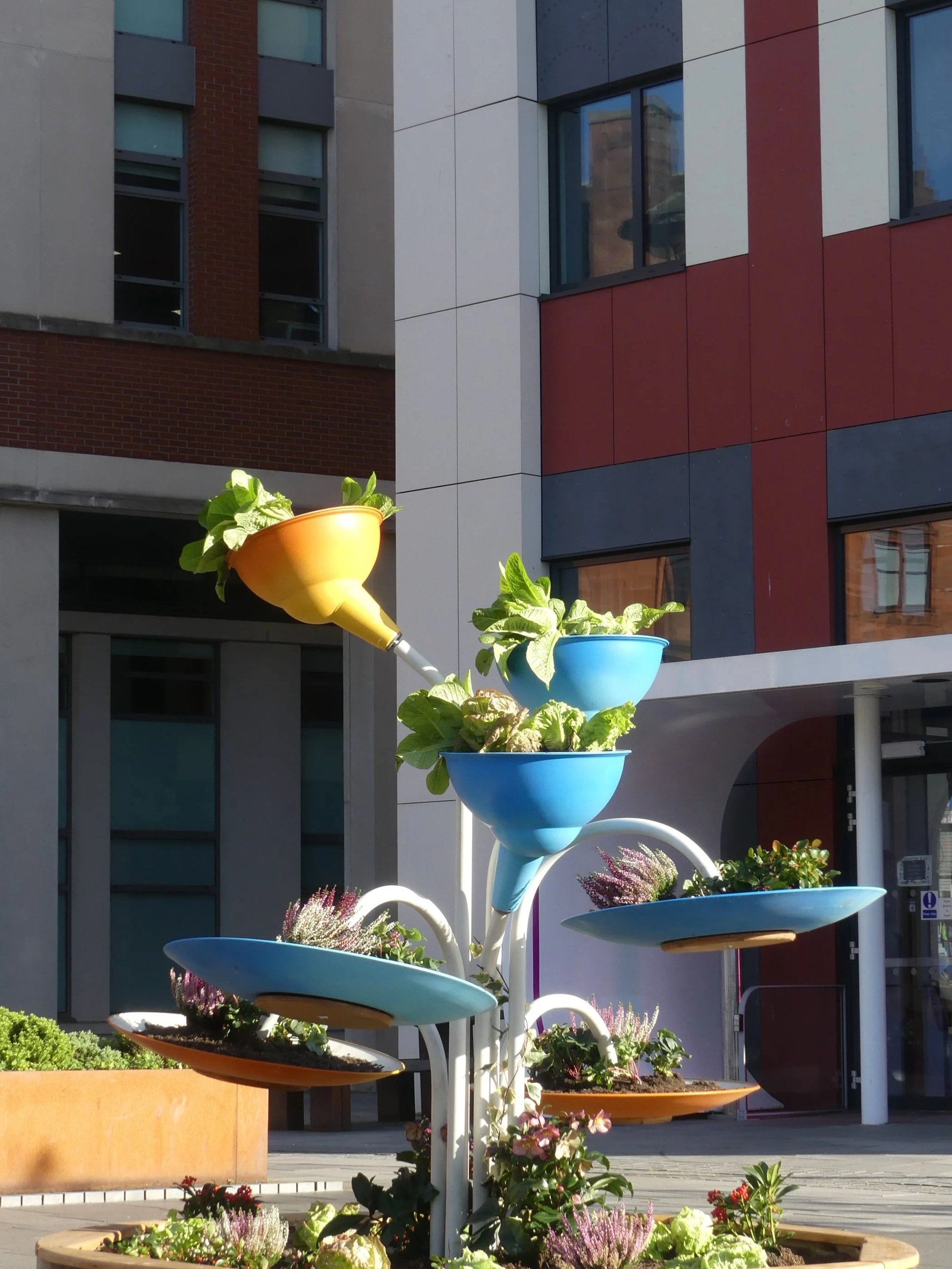 Colorful tiered plant display with various flowers and green plants in containers of different shapes and colors, set outdoors in front of a modern building with large windows and brick and panel exterior.