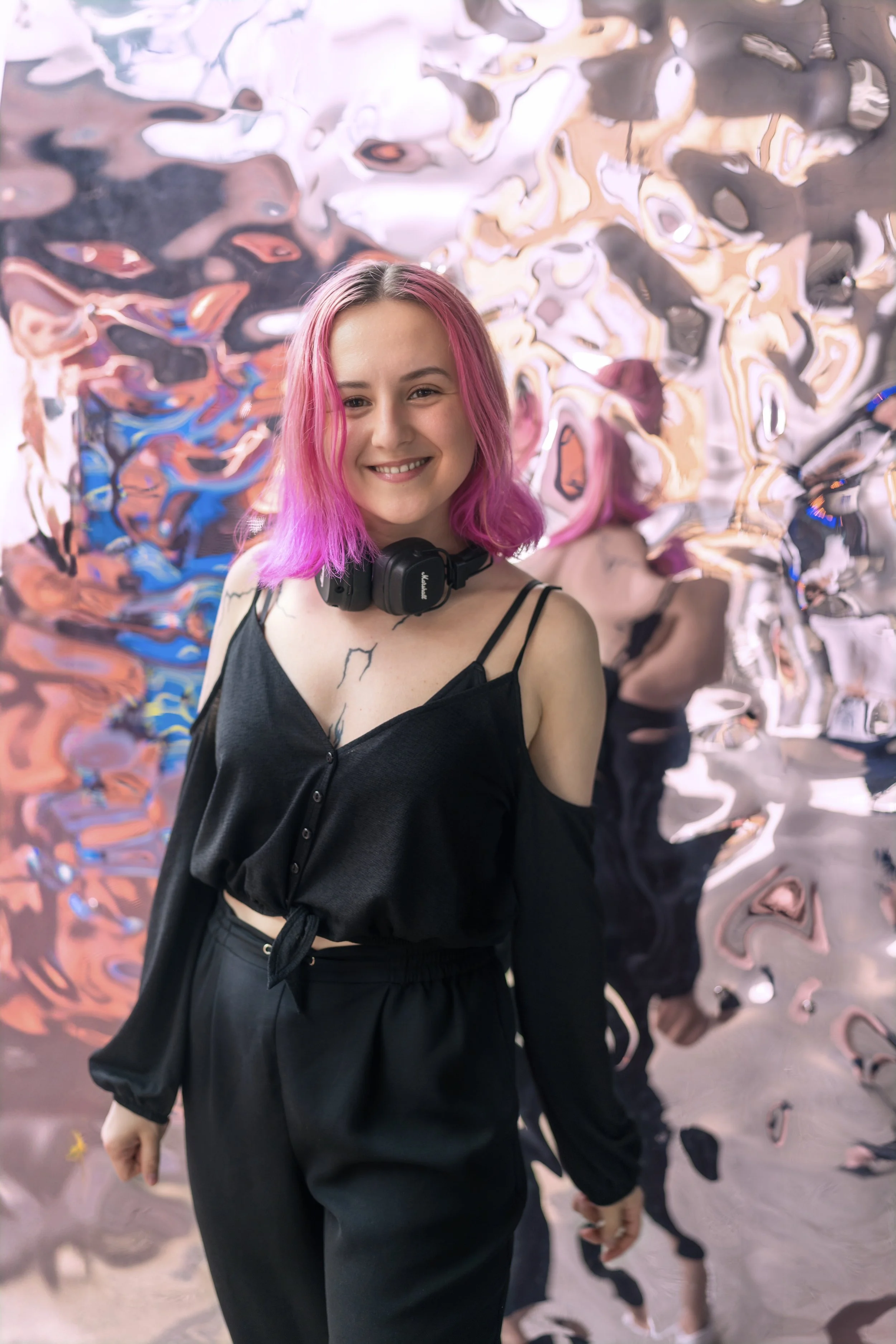 JFOX DJ smiling at the camera, wearing a black off-the-shoulder top and black pants, standing in front of an abstract, distorted reflective background.