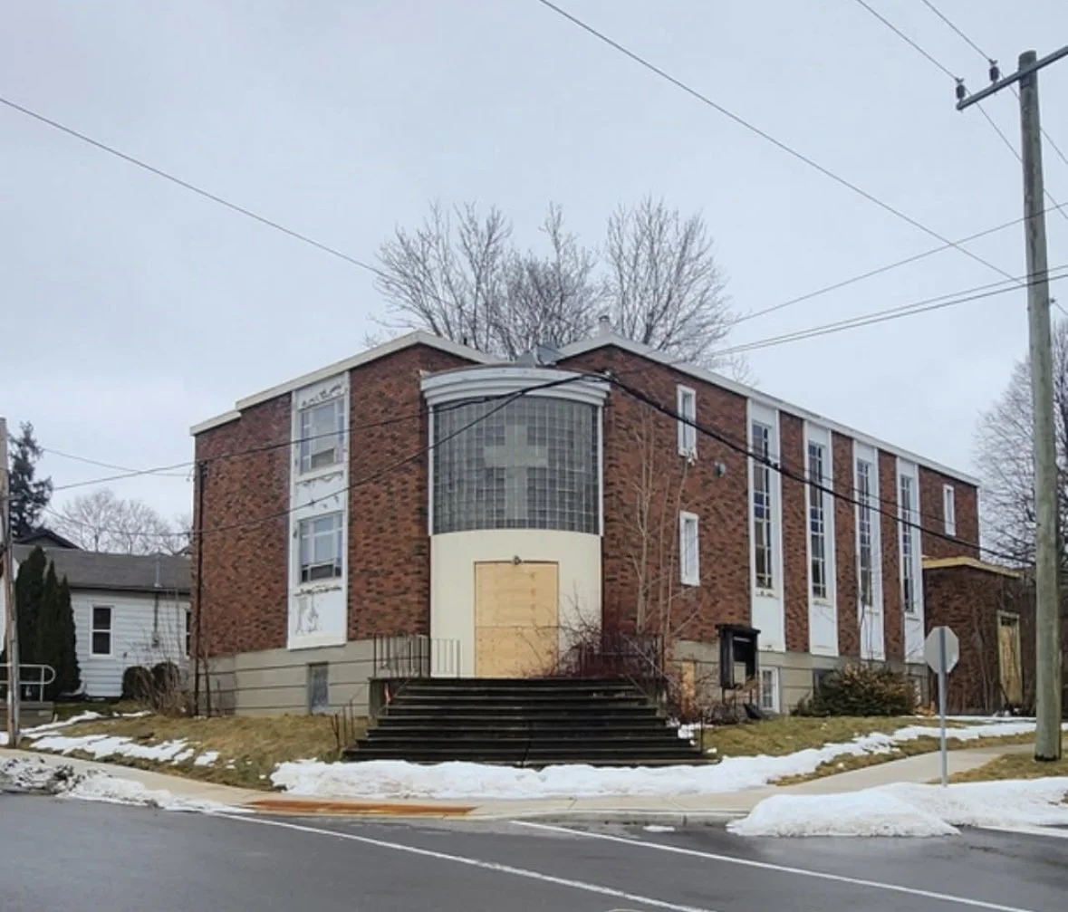 A brick church building with tall vertical windows, a glass block section with a cross, and steps leading up to a boarded-up door, situated on a corner lot with snow on the ground.