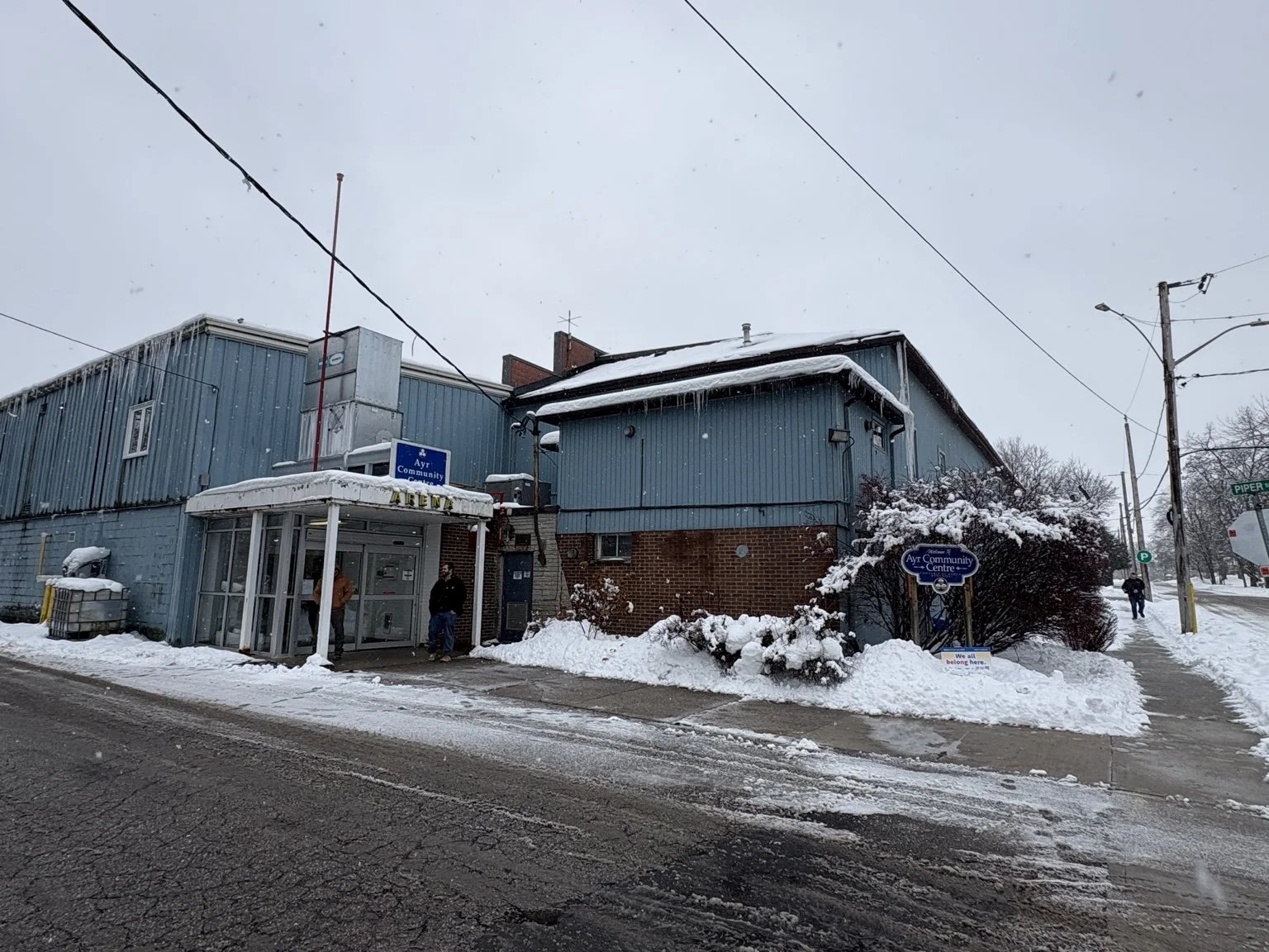 Snow-covered blue and brick community center building with people standing outside in winter, snow on the ground and roof, utility poles, and a sign reading 'Ayr Community Centre' near the sidewalk.
