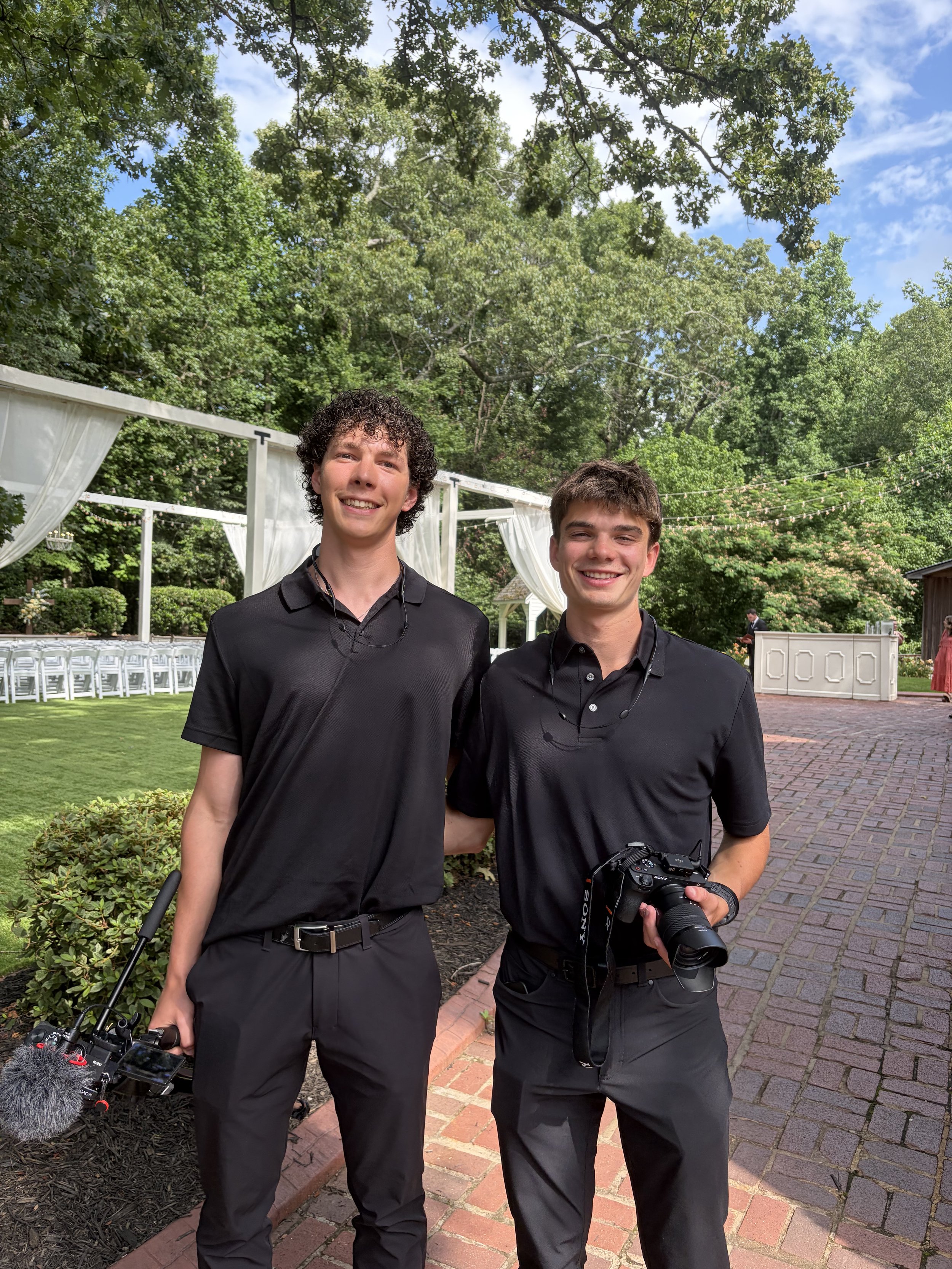 Two young men dressed in black shirts standing outdoors in a garden setting, smiling, with decorative white curtains and greenery in the background.
