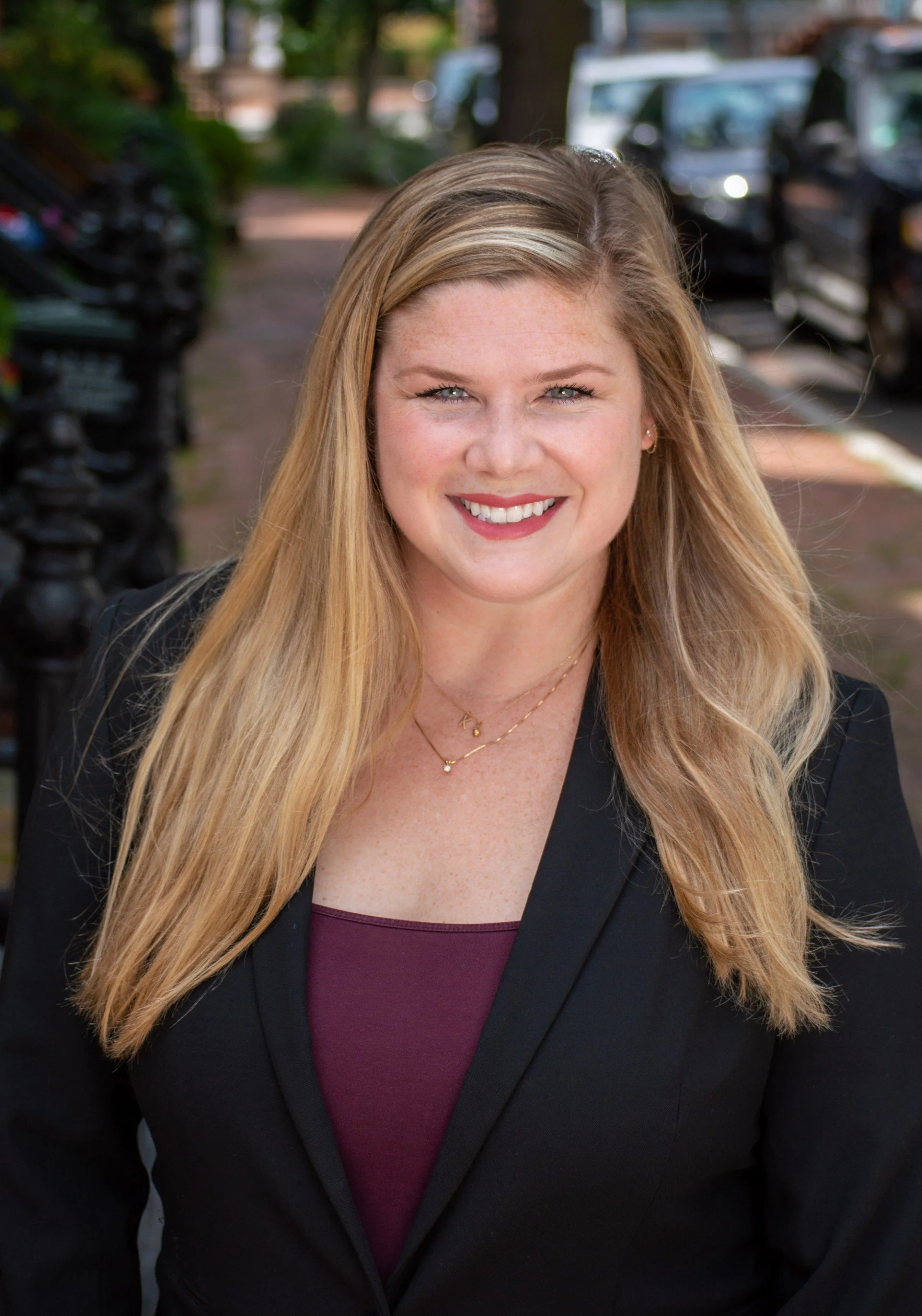 Close-up of a smiling woman with long blonde hair, wearing a black blazer over a maroon top, standing outdoors with blurred cars and trees in the background.