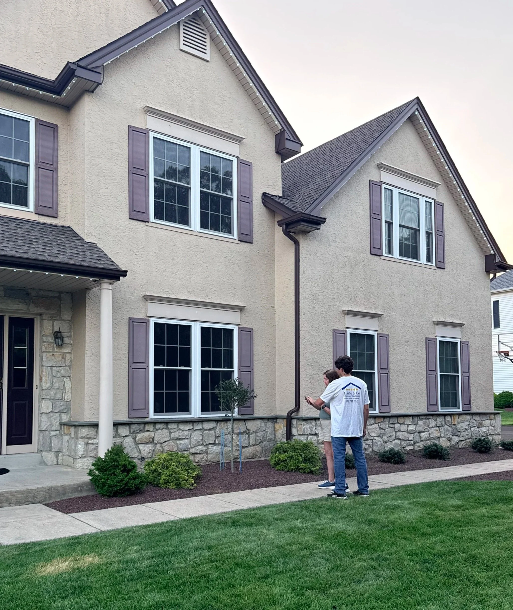 A residential house with beige stucco exterior, gray roof, and purple window shutters. Two people are standing on the sidewalk, looking at a smartphone. The house has a small garden with bushes and a newly planted tree, and the lawn is green.