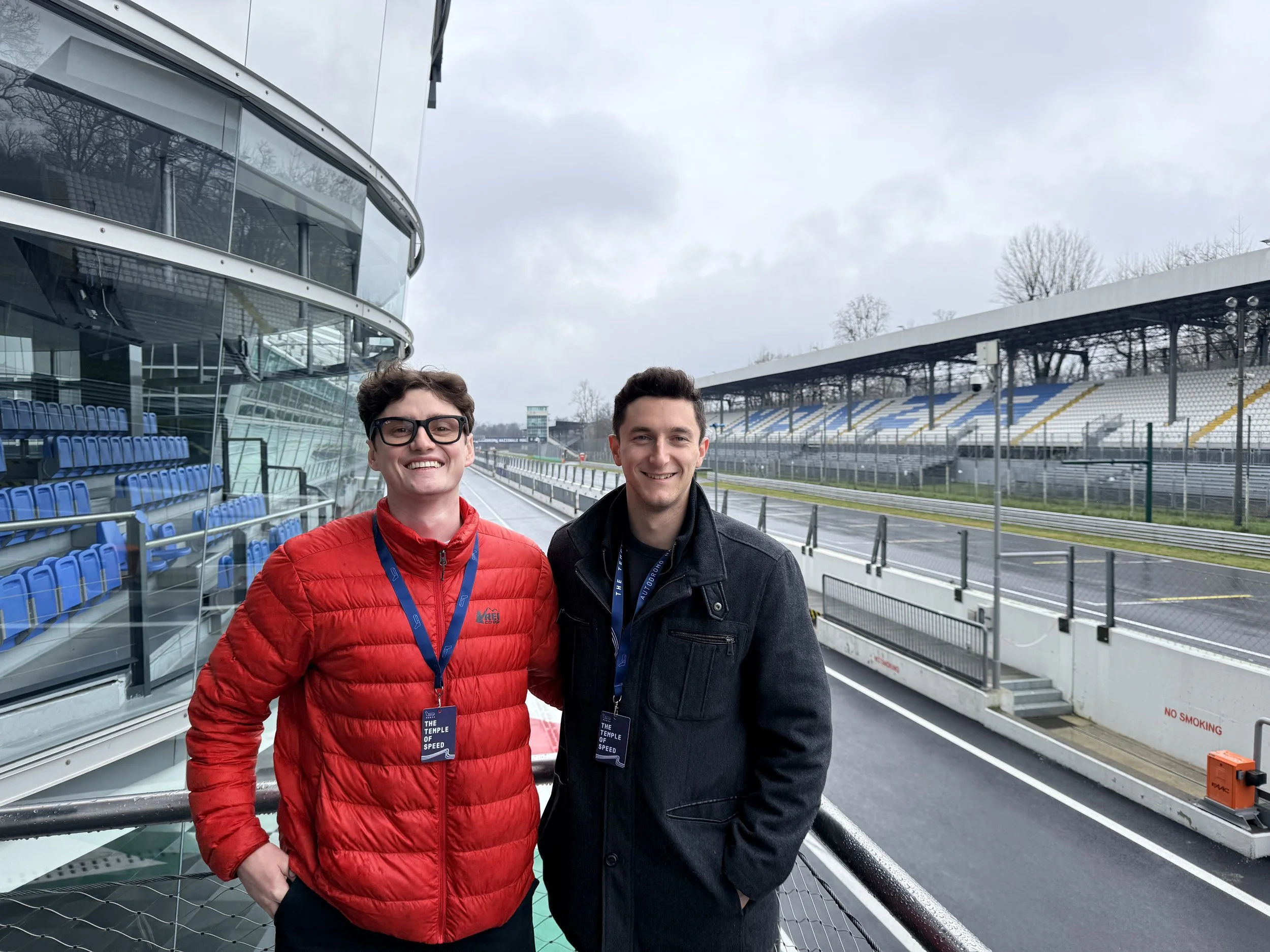 Two young men smiling at a racetrack pit lane, with empty stands, glass-fronted garages, and a cloudy sky behind them.