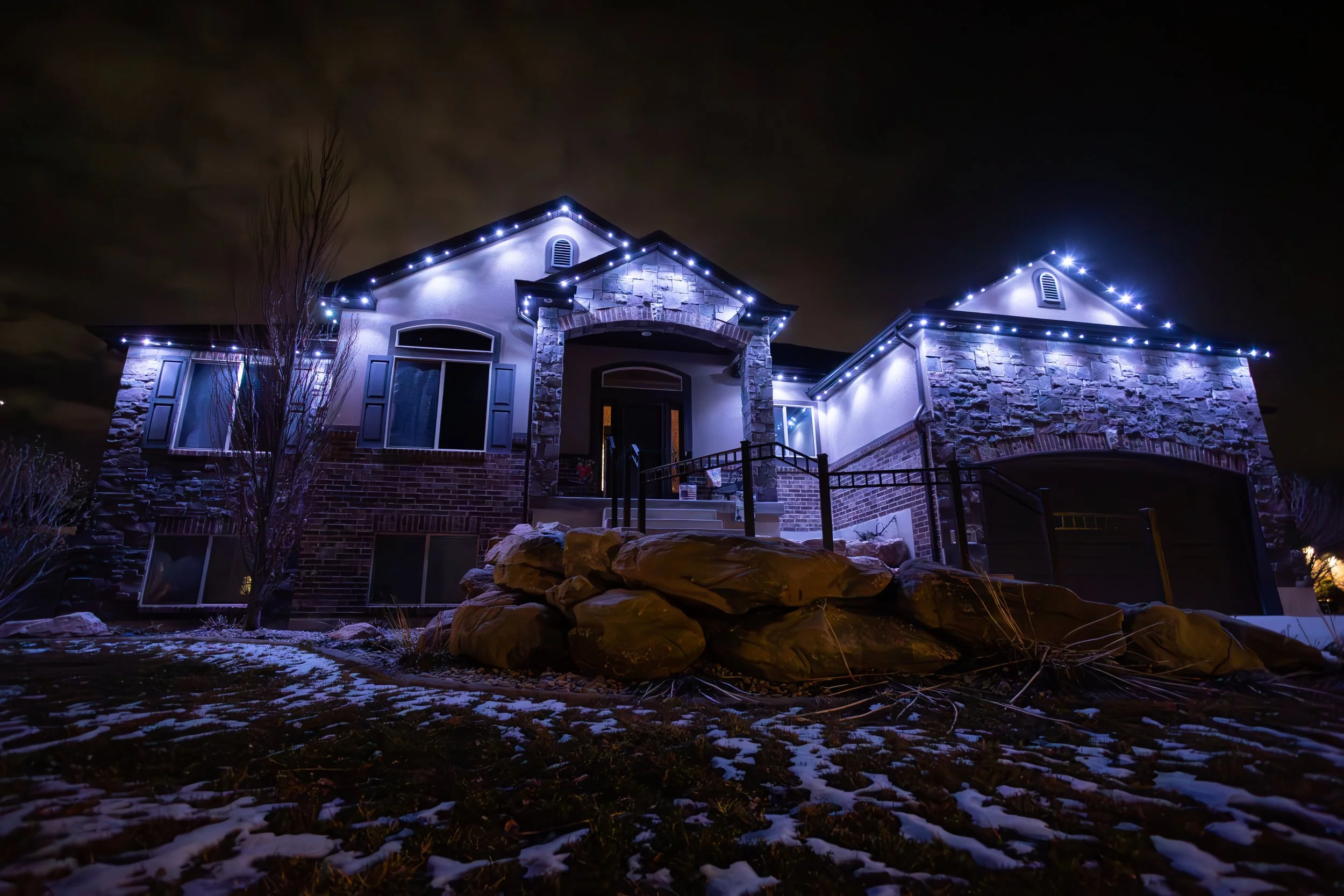 A two-story house decorated with white and blue Christmas lights at night, with a brick and stone exterior, front stairs, and a yard with rocks and some patches of snow.