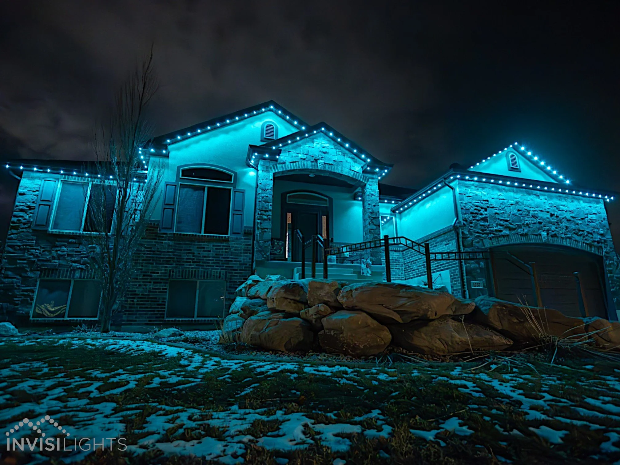 A house decorated with blue Christmas lights at night. The house has brick and stone exterior with a front porch, steps, and large windows. There is a small tree and some snow or ice on the ground.