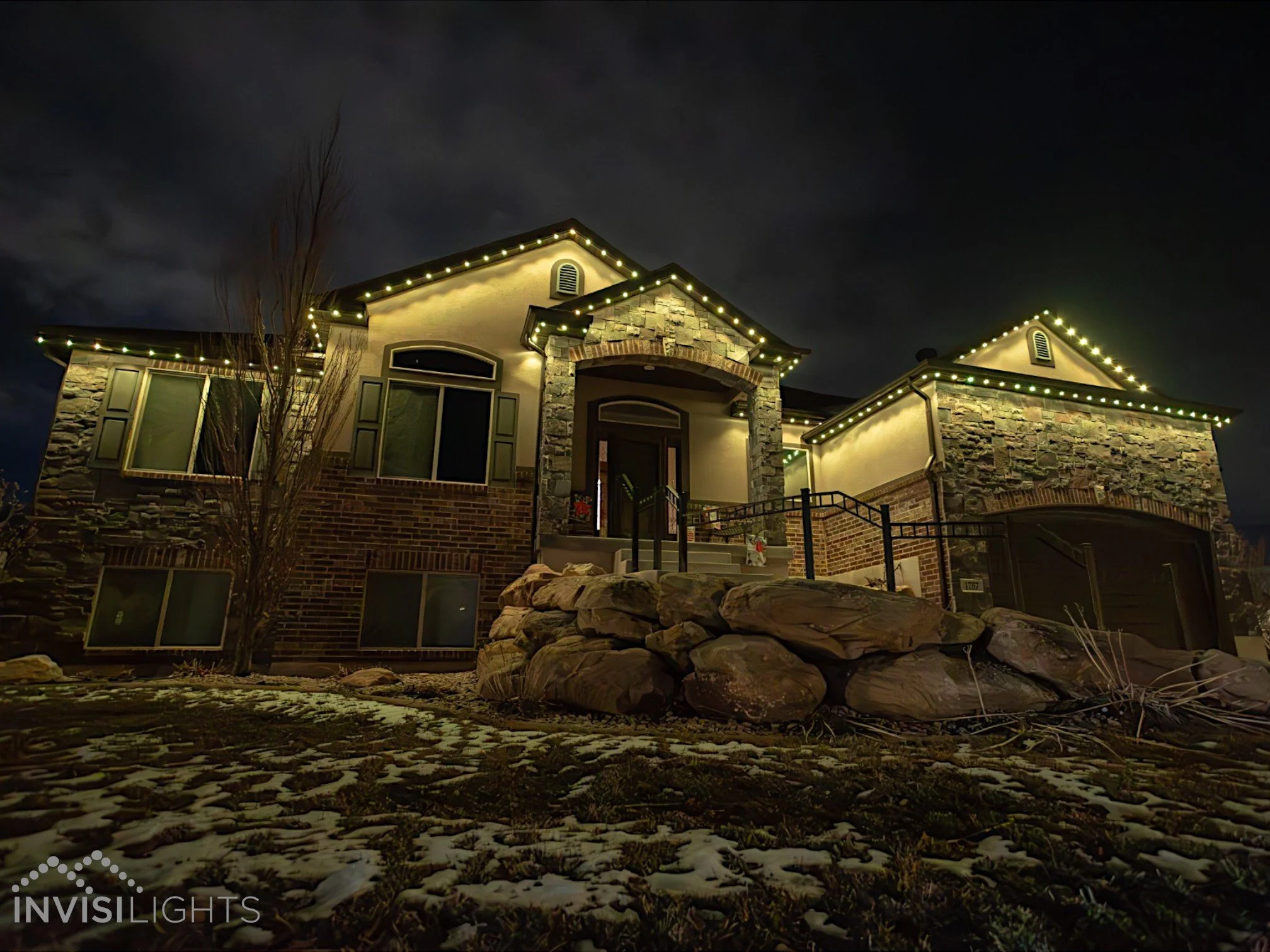 Nighttime photo of a large house decorated with string lights along the roofline. The house has a stone and stucco exterior with a front porch and stairs. There is a leafless tree in front, and the ground has patches of snow. The sky is cloudy.