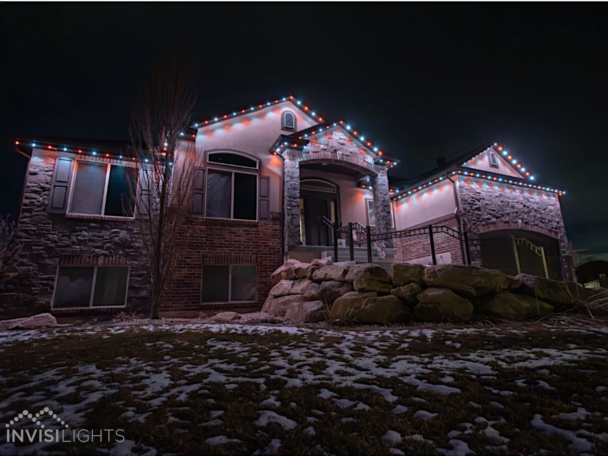 A house decorated with Christmas lights at night. The house has a stone and brick exterior with multiple windows. The roofline is lined with red and white Christmas lights, and the sky is dark.