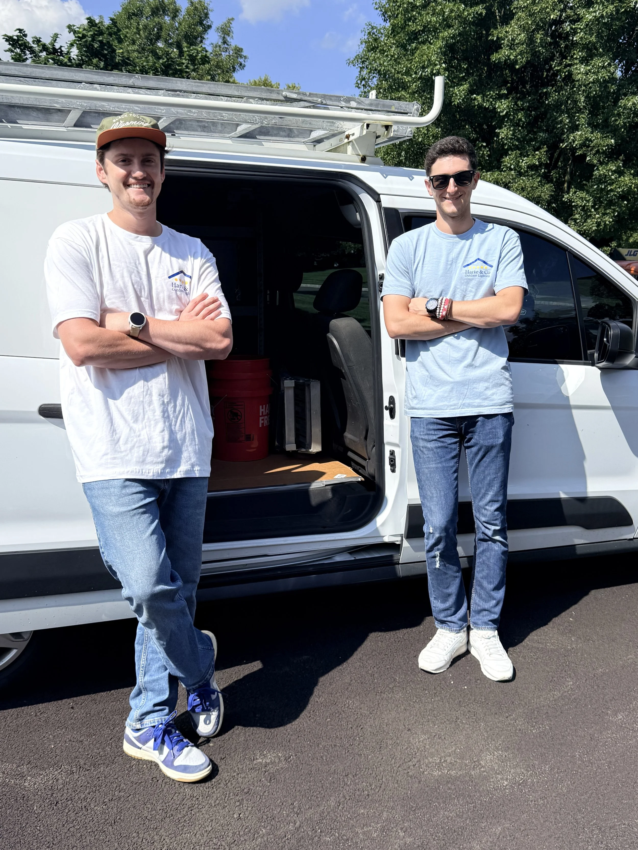 Two young men standing in front of a white work van with open sliding door, outdoors on a sunny day, both smiling and crossing their arms.