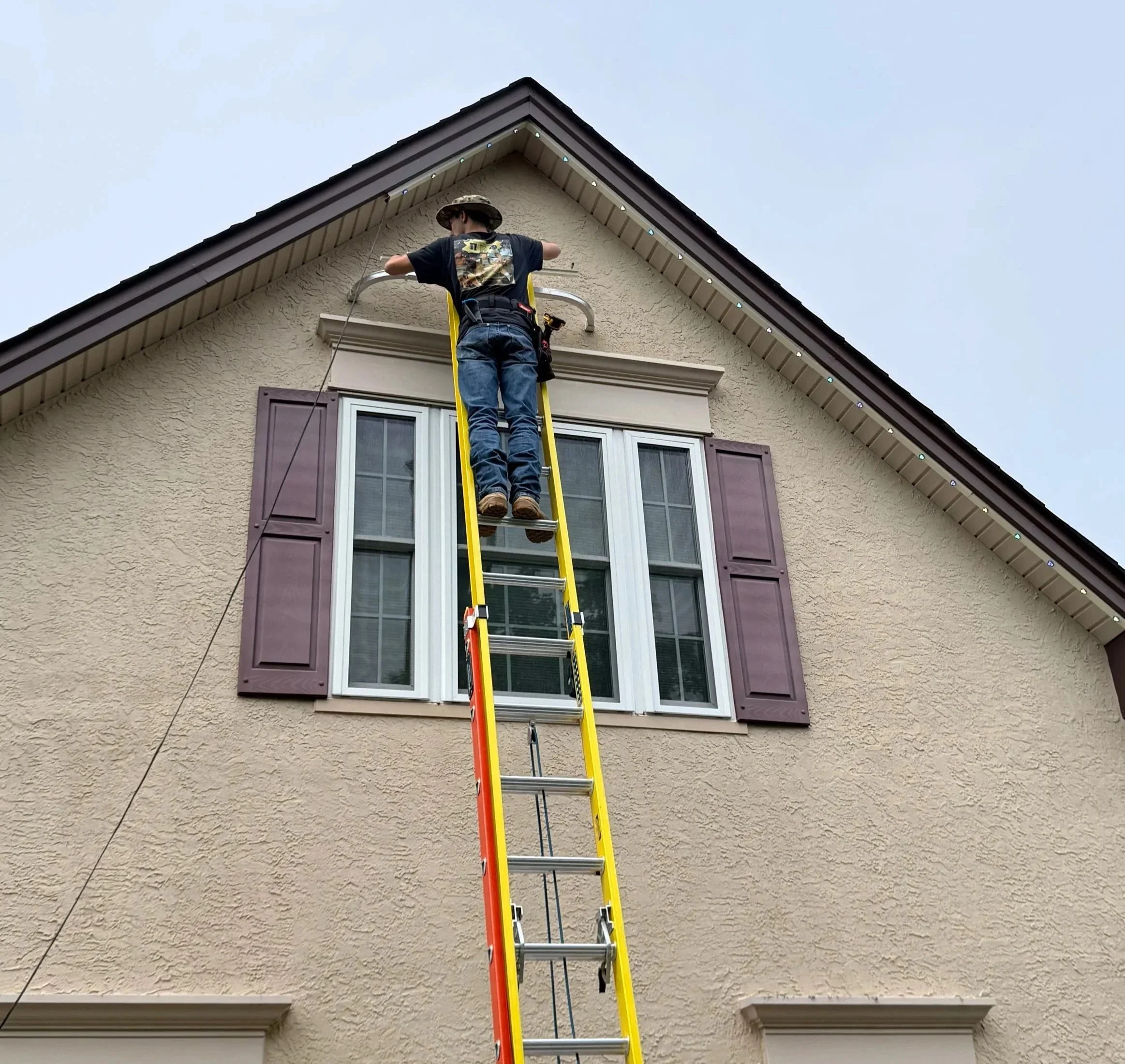 A person wearing a camouflage hat and dark clothing is standing on a yellow ladder while working on the exterior of a house, possibly installing or repairing a gutter or fascia, against a background of a clear sky.