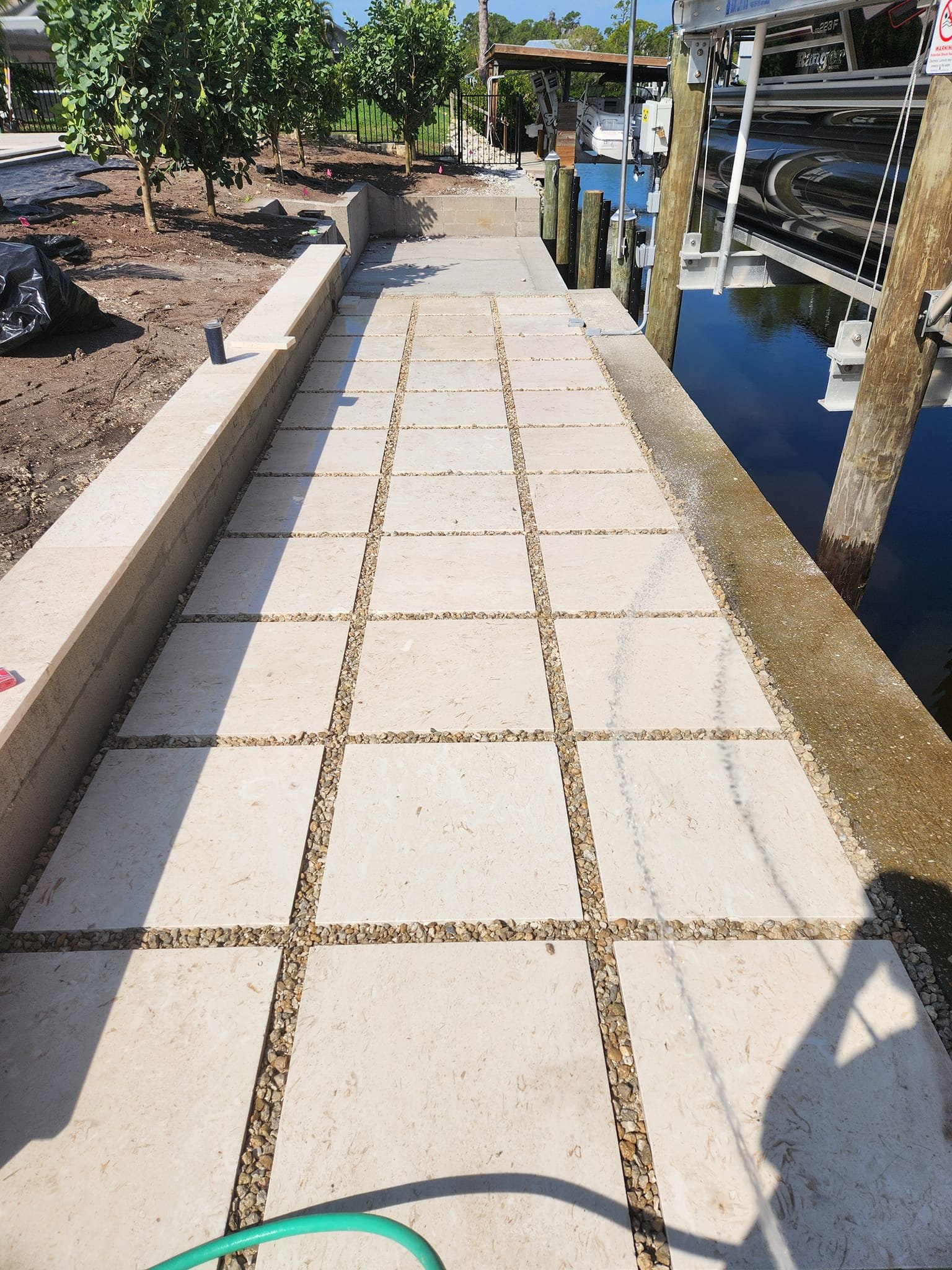 A newly paved walkway with large rectangular concrete pavers and gravel fillers, adjacent to a waterway with a boat lift, in a backyard with trees and landscaping in progress.