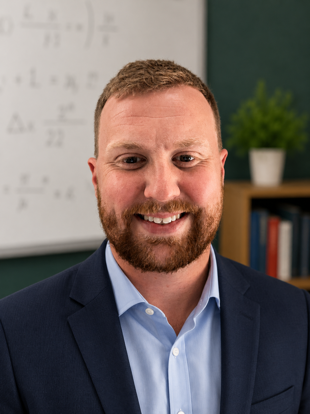 A smiling man with a beard and short hair, wearing a dark suit jacket and light blue shirt, standing in an office with a whiteboard and bookshelf with a potted plant in the background.