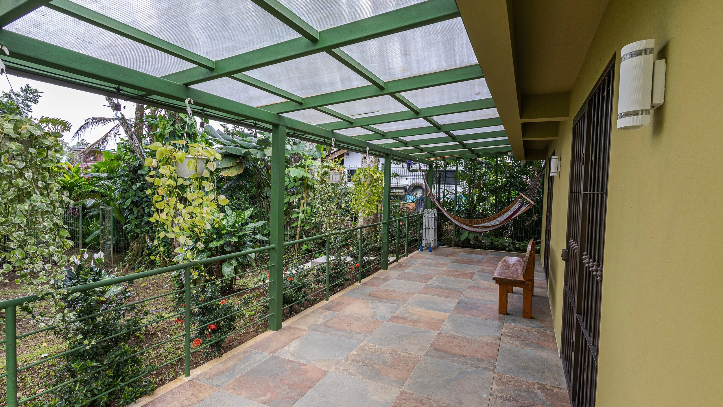 A covered patio with tiled flooring, yellow wall with barred window, and a wooden bench. The patio overlooks a lush garden with various plants and trees, and a hammock is hanging between the plants.