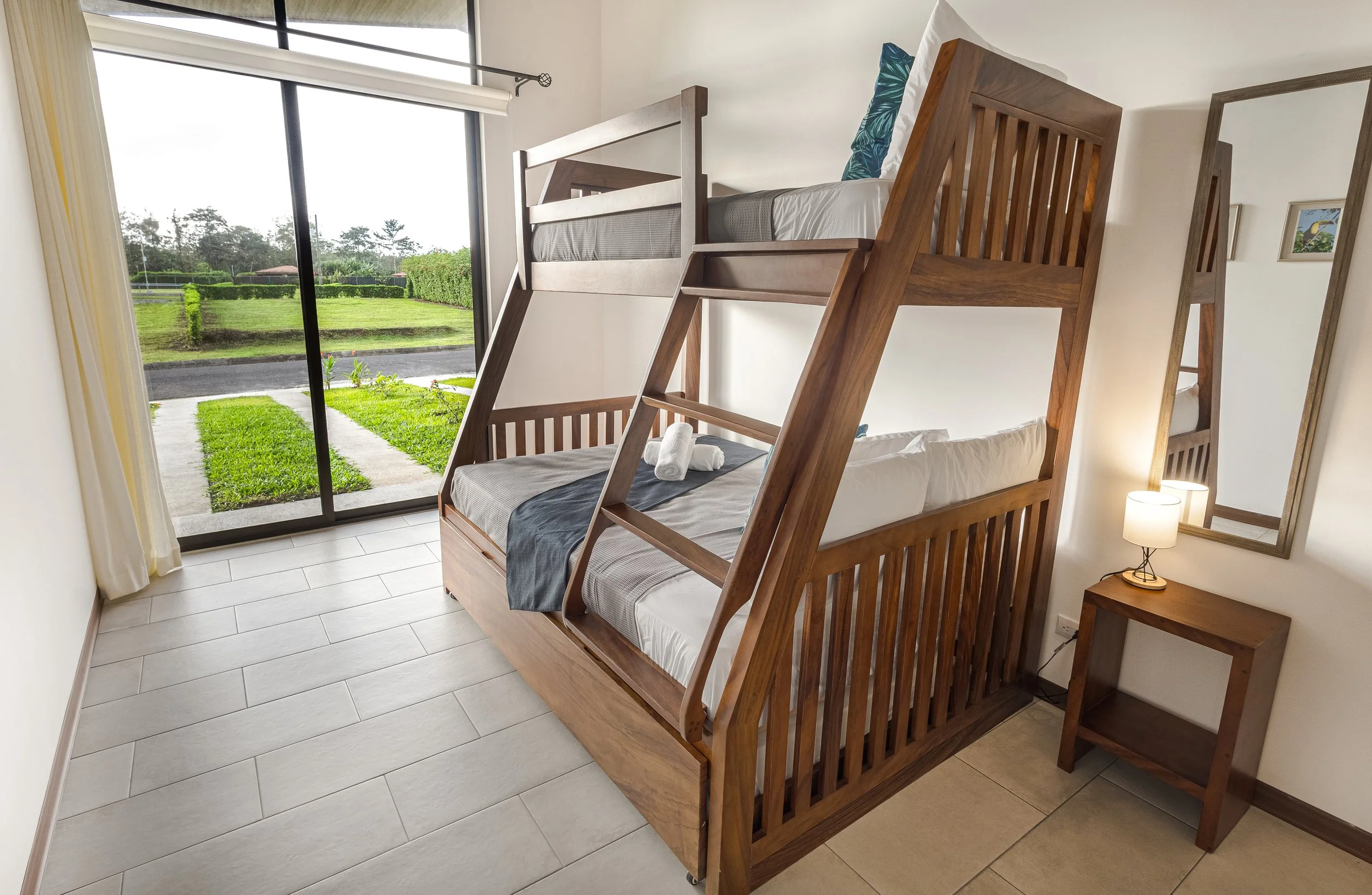 A bedroom with a wooden bunk bed, pillows, and towels folded on the lower bed, next to a nightstand with a lamp, large sliding glass door with curtains open showing a garden outside.