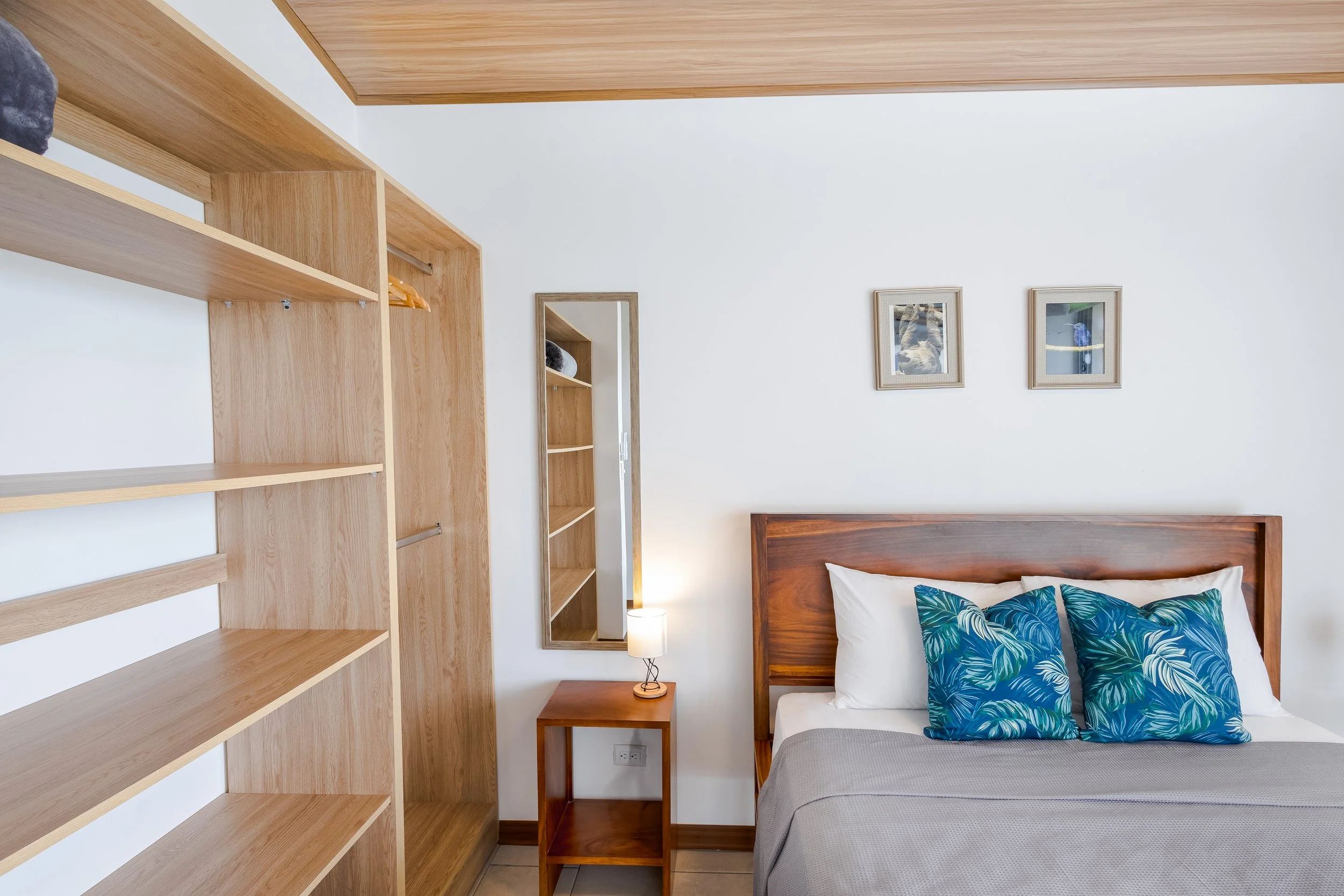 A bedroom with a wooden headboard, two blue tropical-themed pillows, a small wooden nightstand with a lamp, a white wall with framed pictures, open wooden shelving, and a mirror.