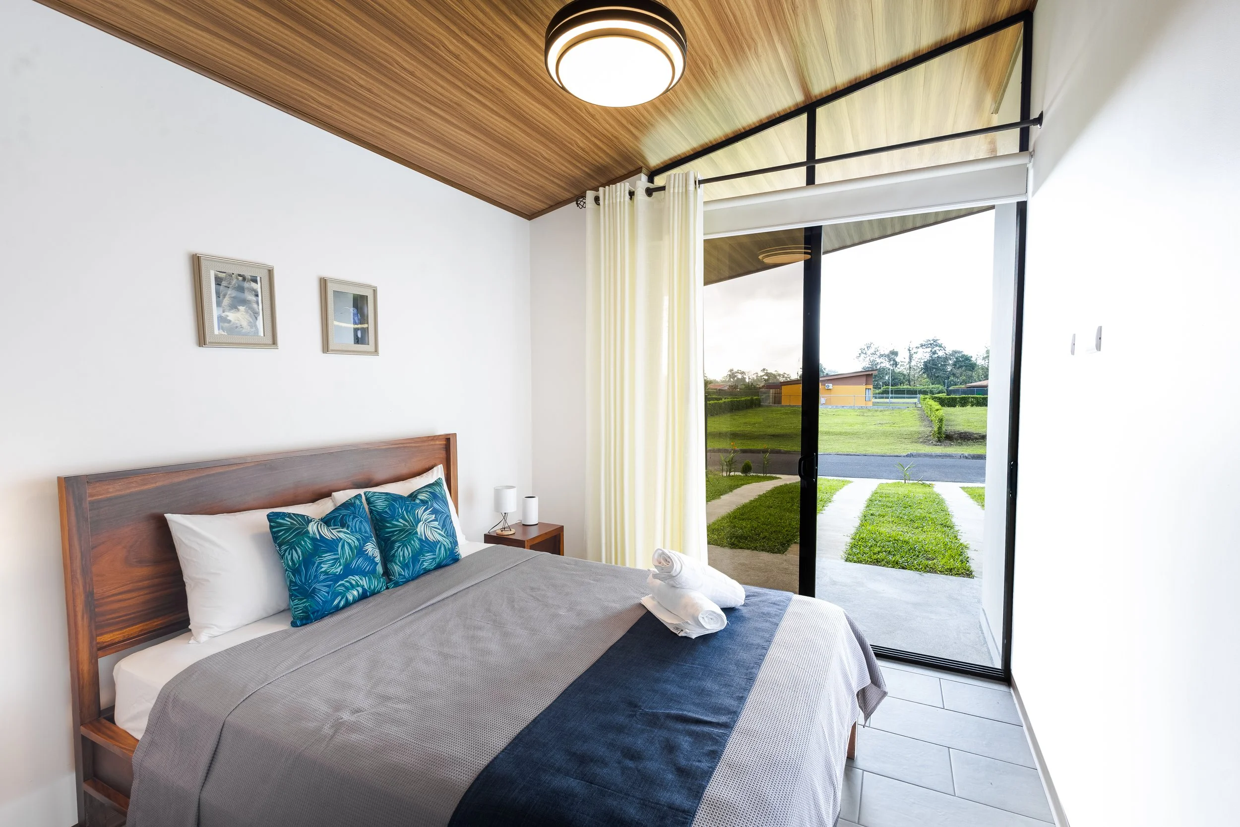 A bedroom with a large glass door opening to a green yard. There is a bed with blue and white pillows, a gray blanket, and towels on top. The room has a wooden ceiling with a circular light fixture, and white walls with two framed pictures.