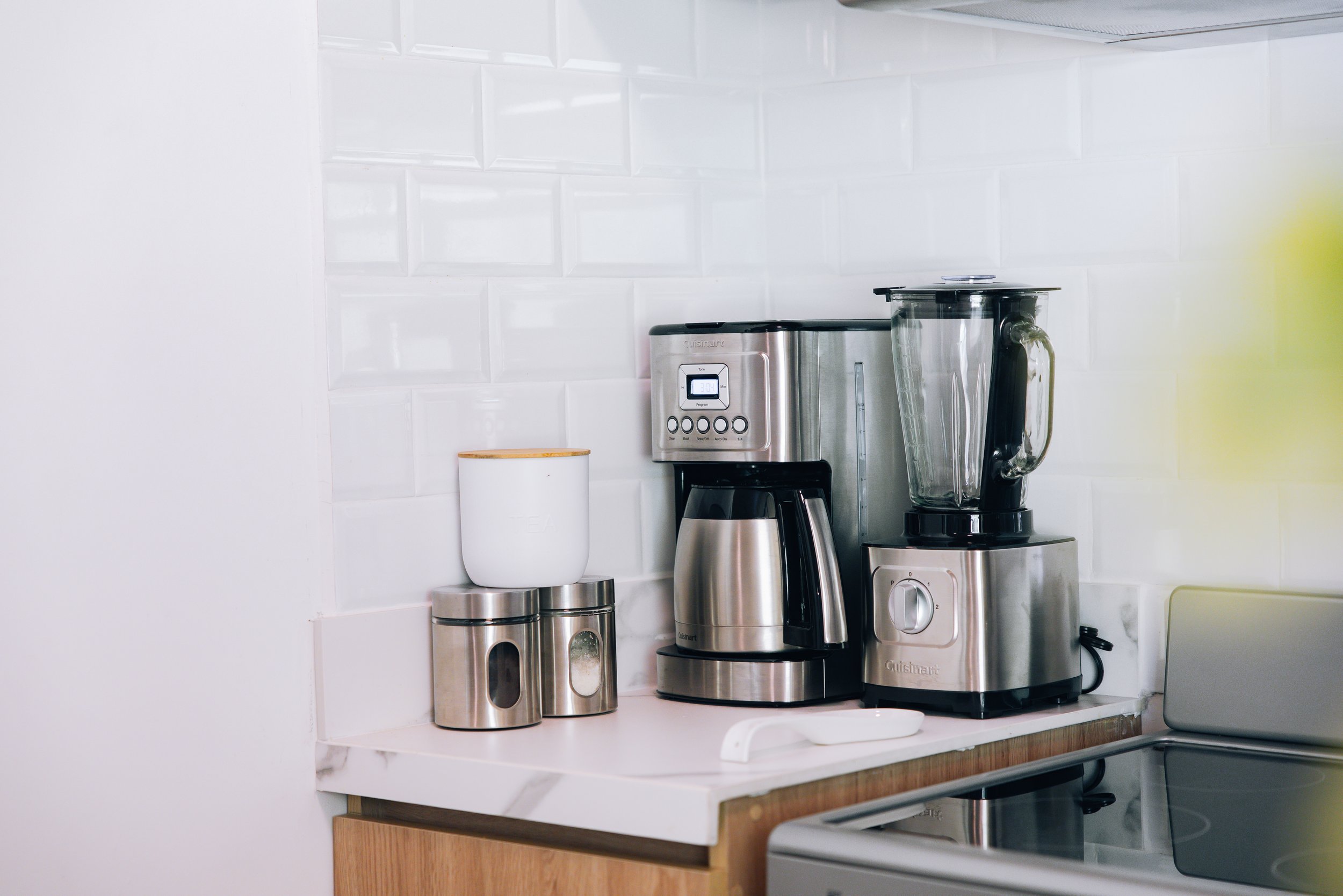 Kitchen countertop with a blend of modern and traditional appliances, including a coffee maker, a blender, and two stainless steel canisters, against a white subway tile backsplash.