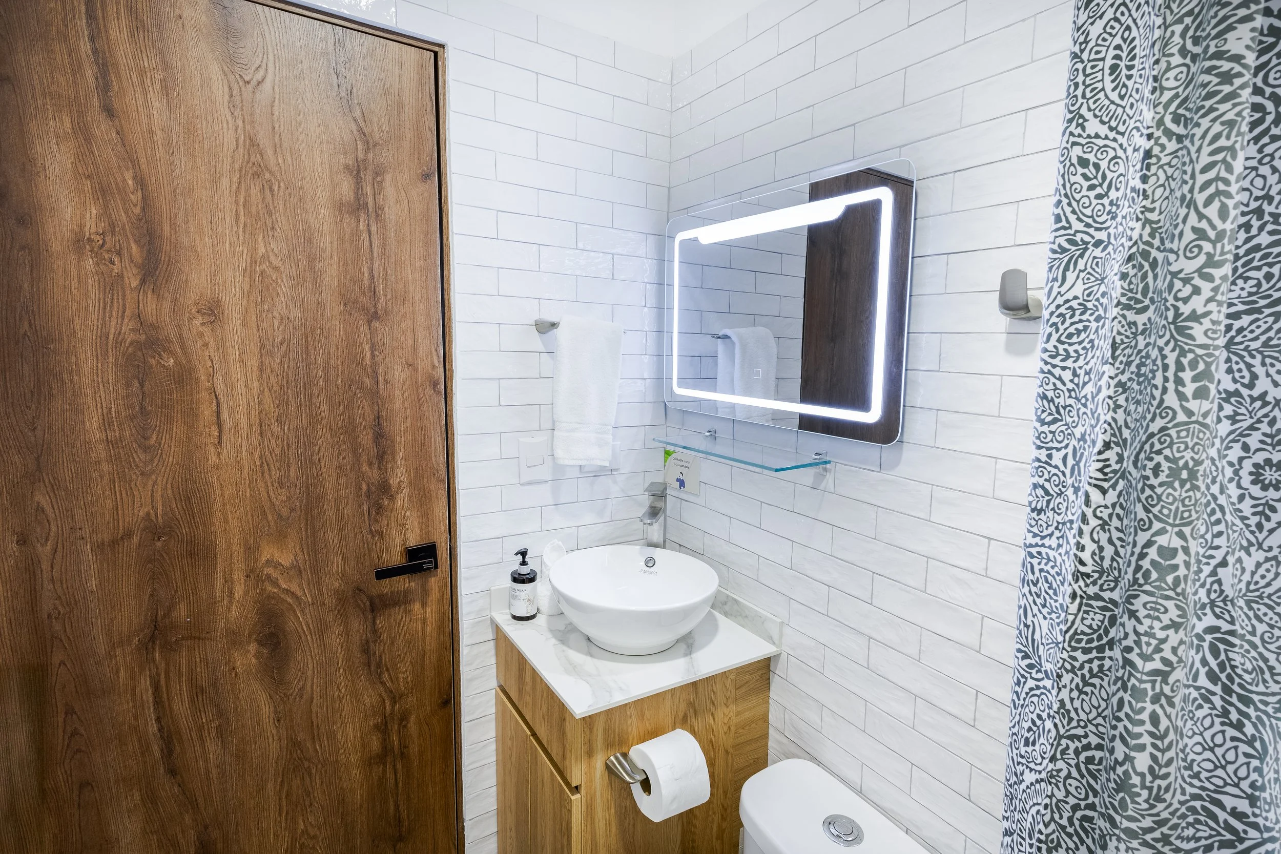 A modern bathroom with white brick tiles, wooden accents, a mirror with LED lighting, and a patterned shower curtain.