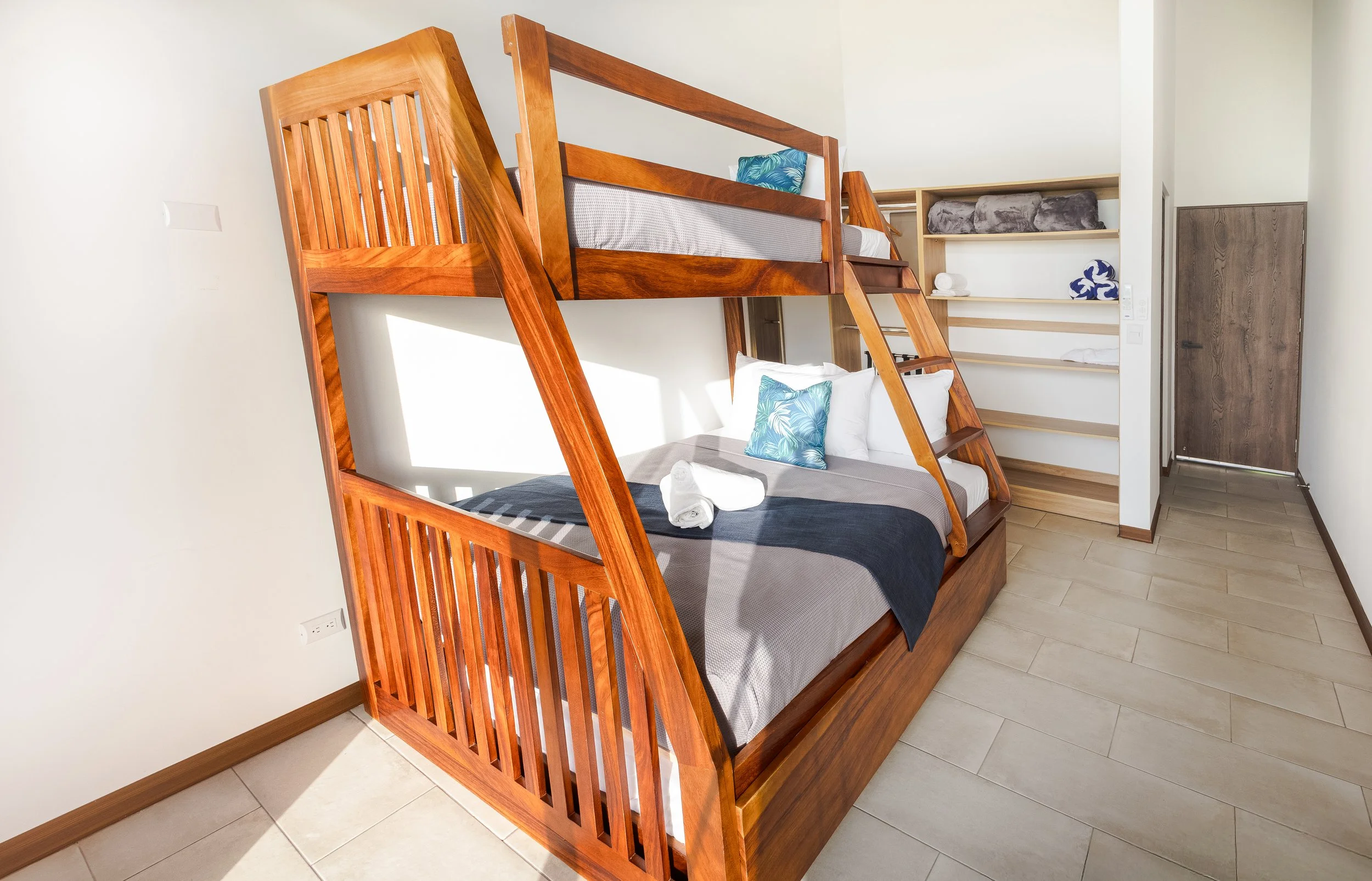Room with wooden bunk beds, a pillow with a blue and green tropical pattern, a wall shelf with rolled towels, a blanket, and a door.