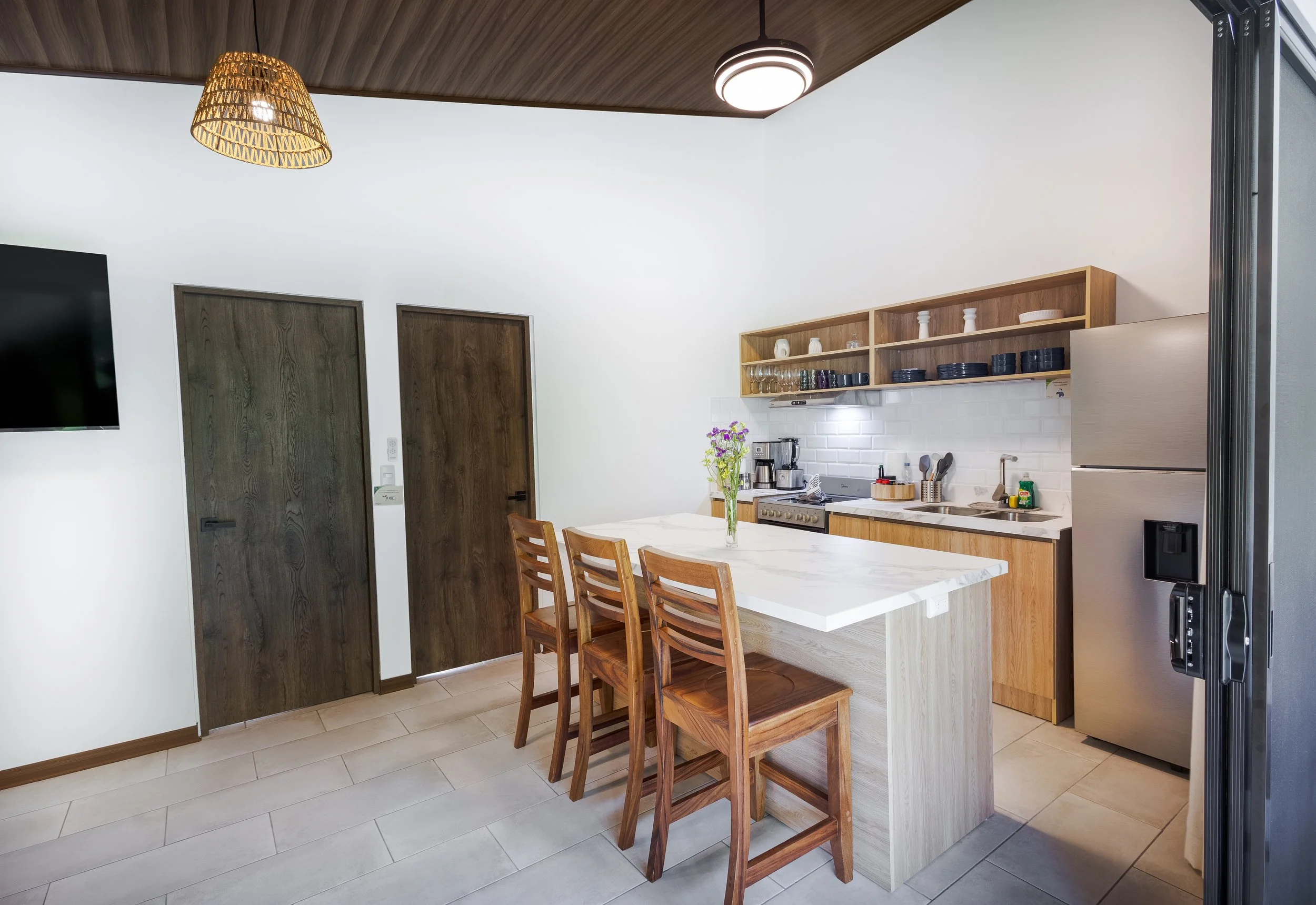 Modern kitchen with wooden cabinets, white countertop, stainless steel appliances, a bunch of purple flowers on the table, and a set of four wooden chairs.