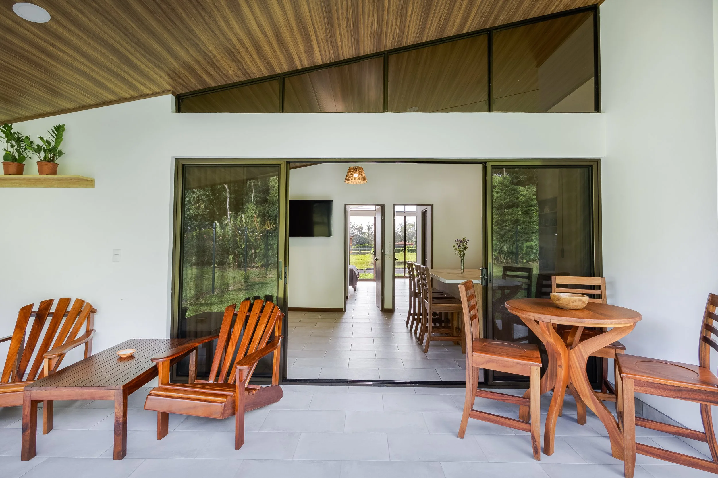 Interior of a modern house with large glass sliding doors, wooden dining tables and chairs, a wall-mounted TV, and outdoor view of trees and grass.