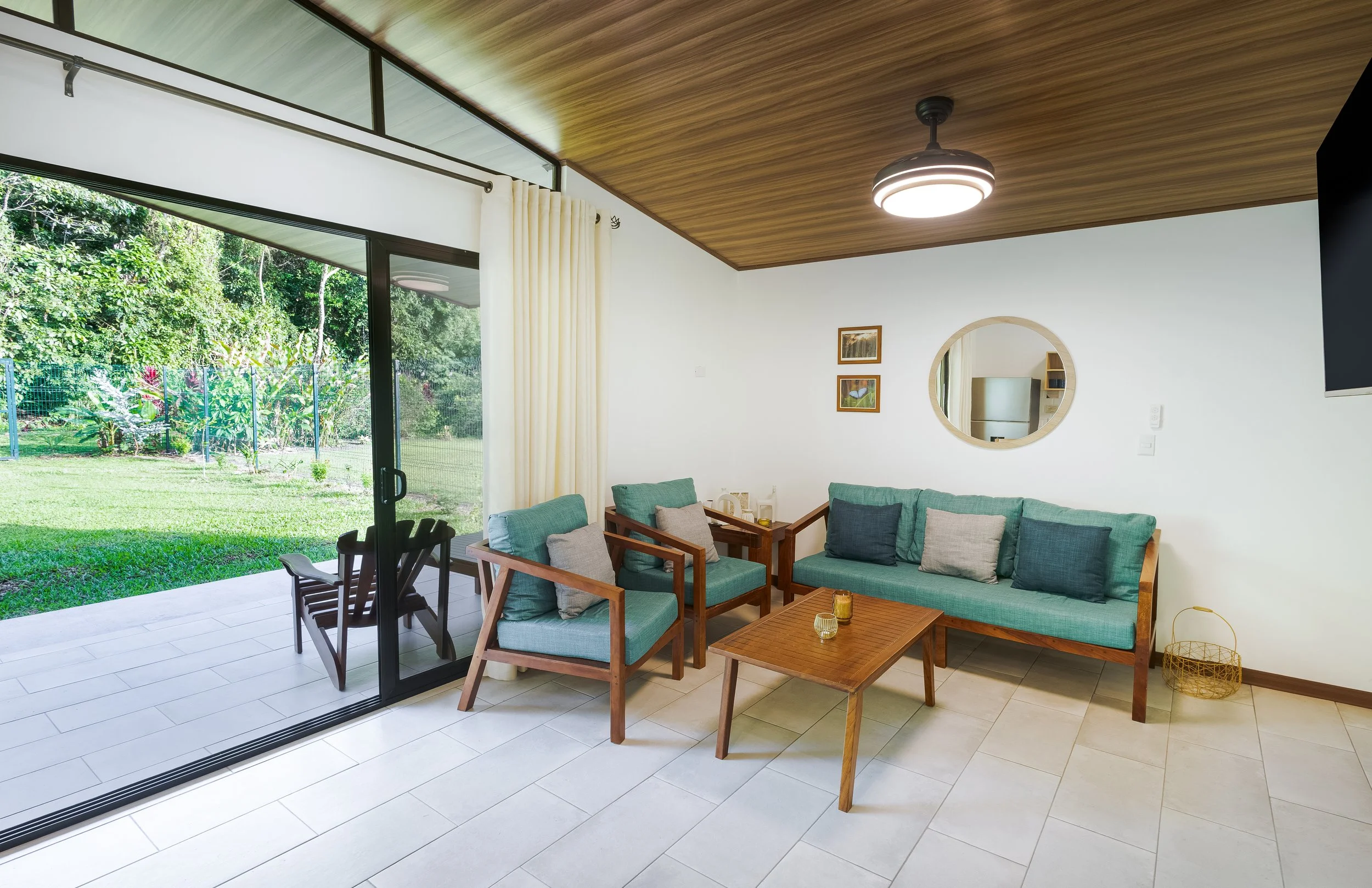 Living room with large glass sliding door leading to a backyard, wooden ceiling, modern ceiling light, circular mirror on white wall, teal couch with pillows, wooden coffee table, and outdoor chairs.