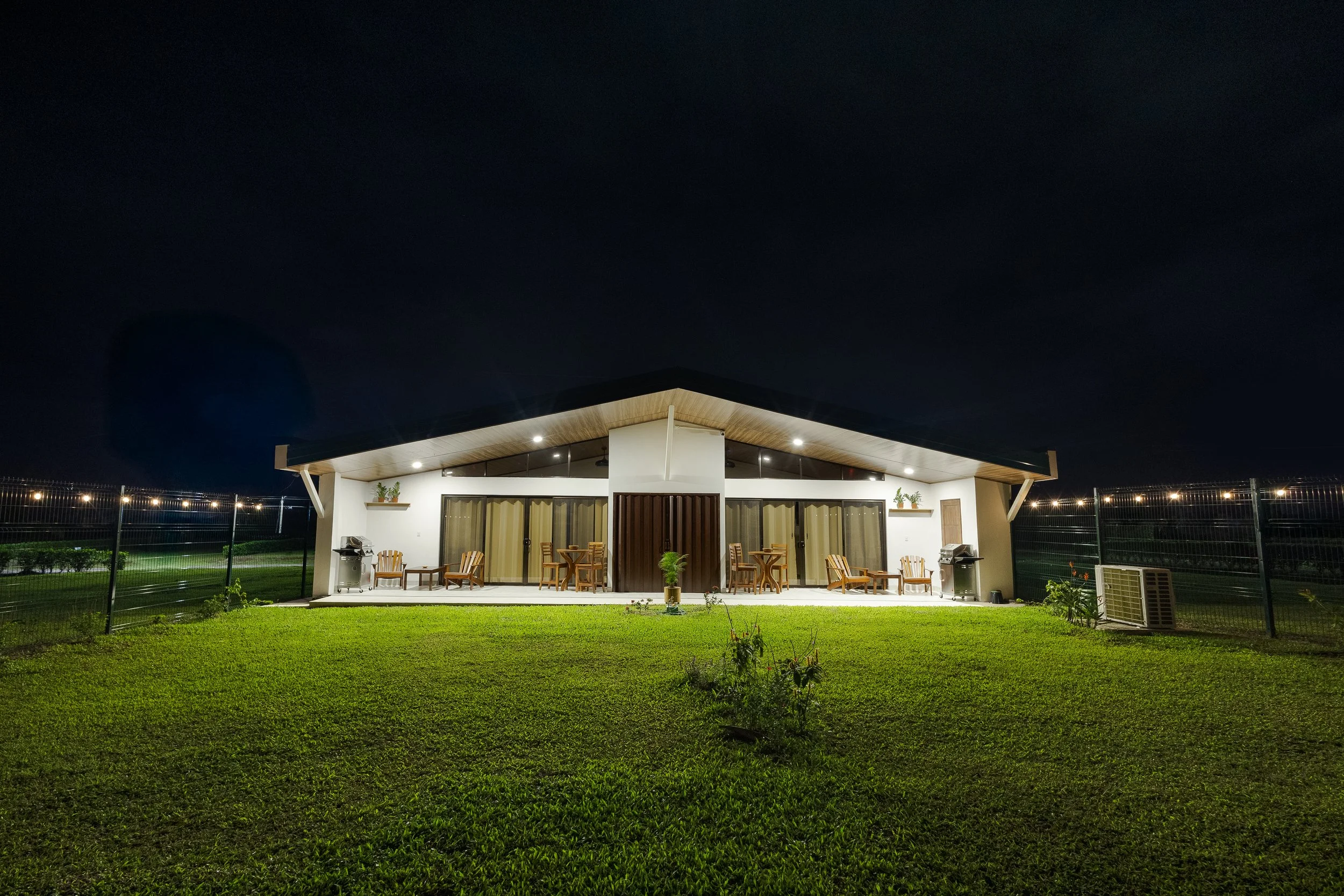 A house with a spacious porch illuminated at night, featuring outdoor furniture, potted plants, and a green lawn enclosed by a metal fence, with a dark sky overhead.
