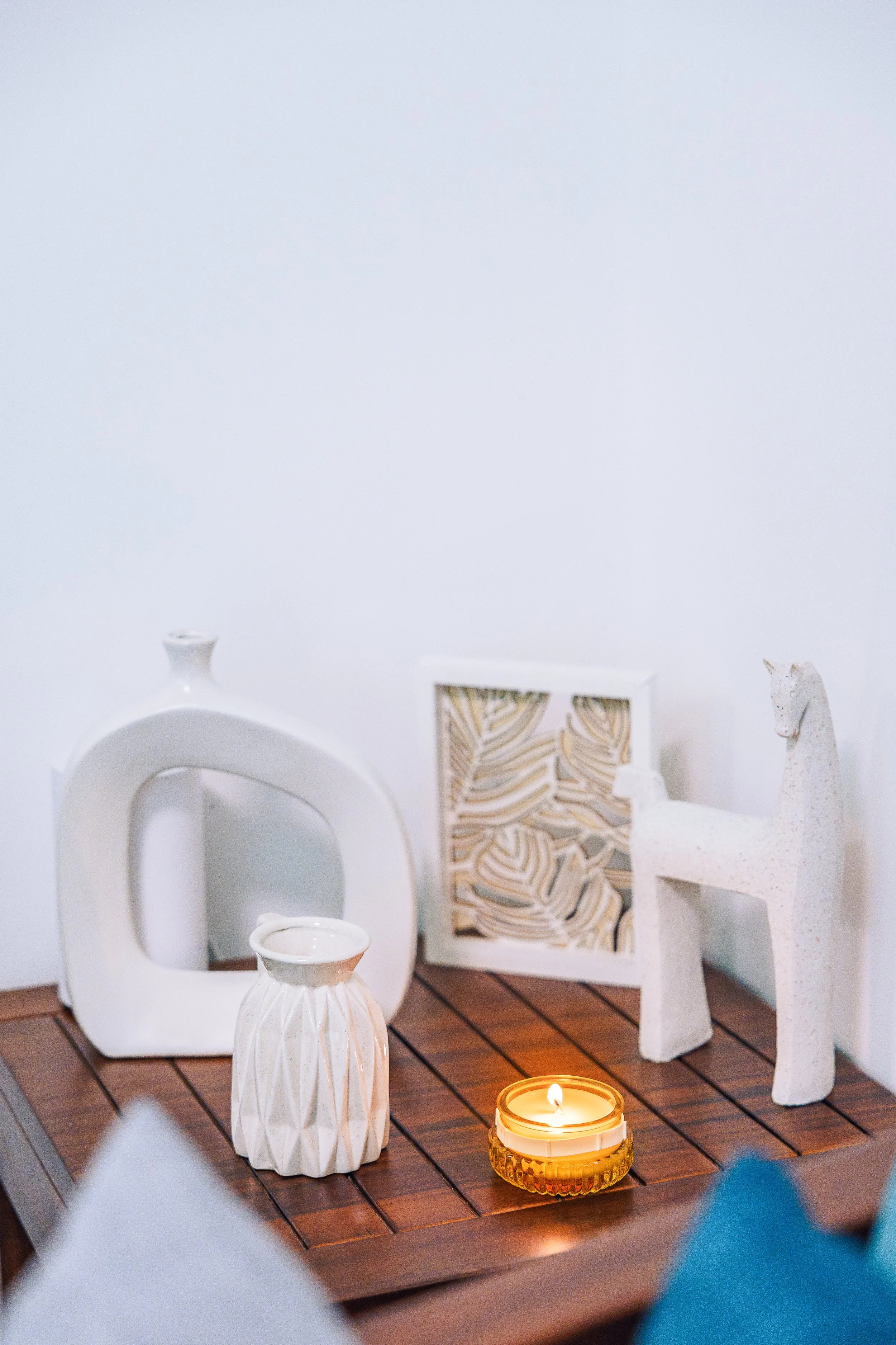 Decorative objects on a wooden table including a white ceramic vase, a tall white textured vase, a small white cup, a white geometric sculpture of a goat, and a lit candle in a small amber glass container. Behind these items, there are framed abstrac