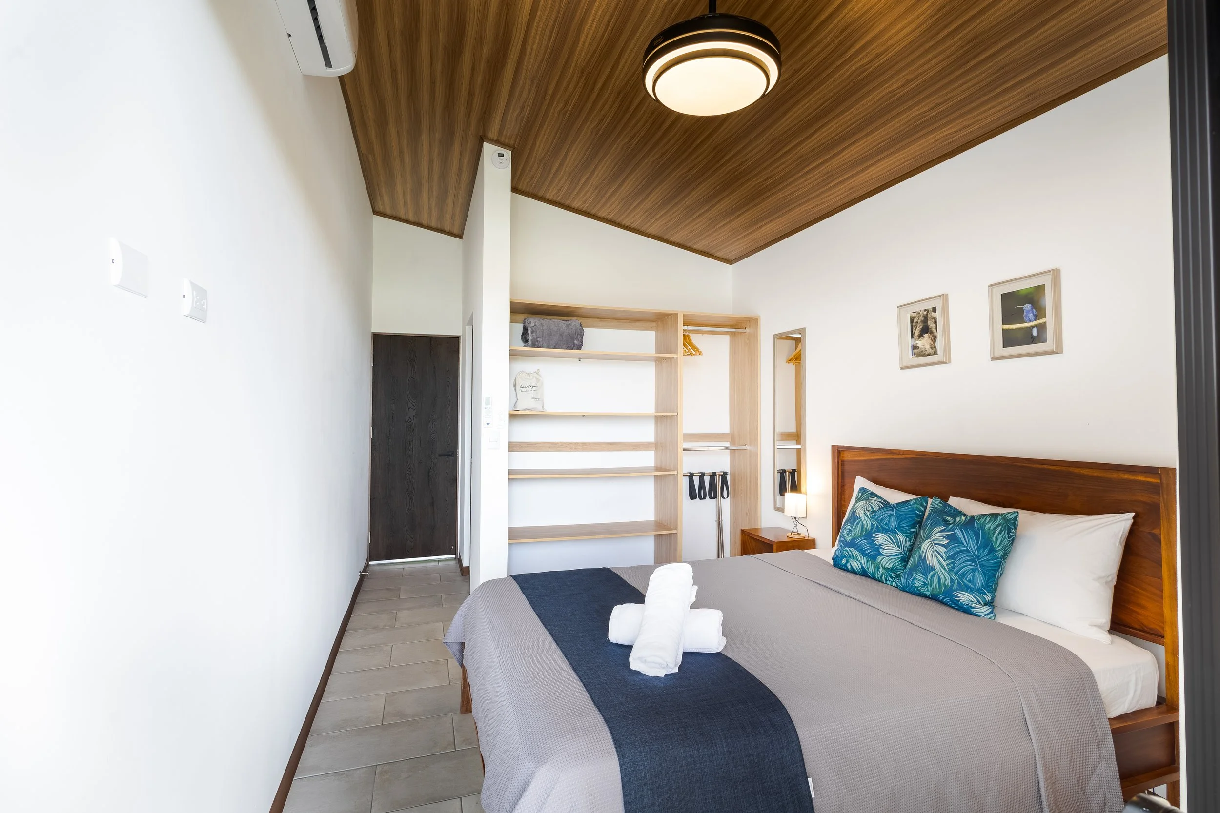 A bedroom with a bed that has white and grey bedding, decorative pillows with blue and white leaf patterns, and towels on top. The room features a wooden headboard, a bedside table with a lamp, and a wooden ceiling. There are framed pictures of birds