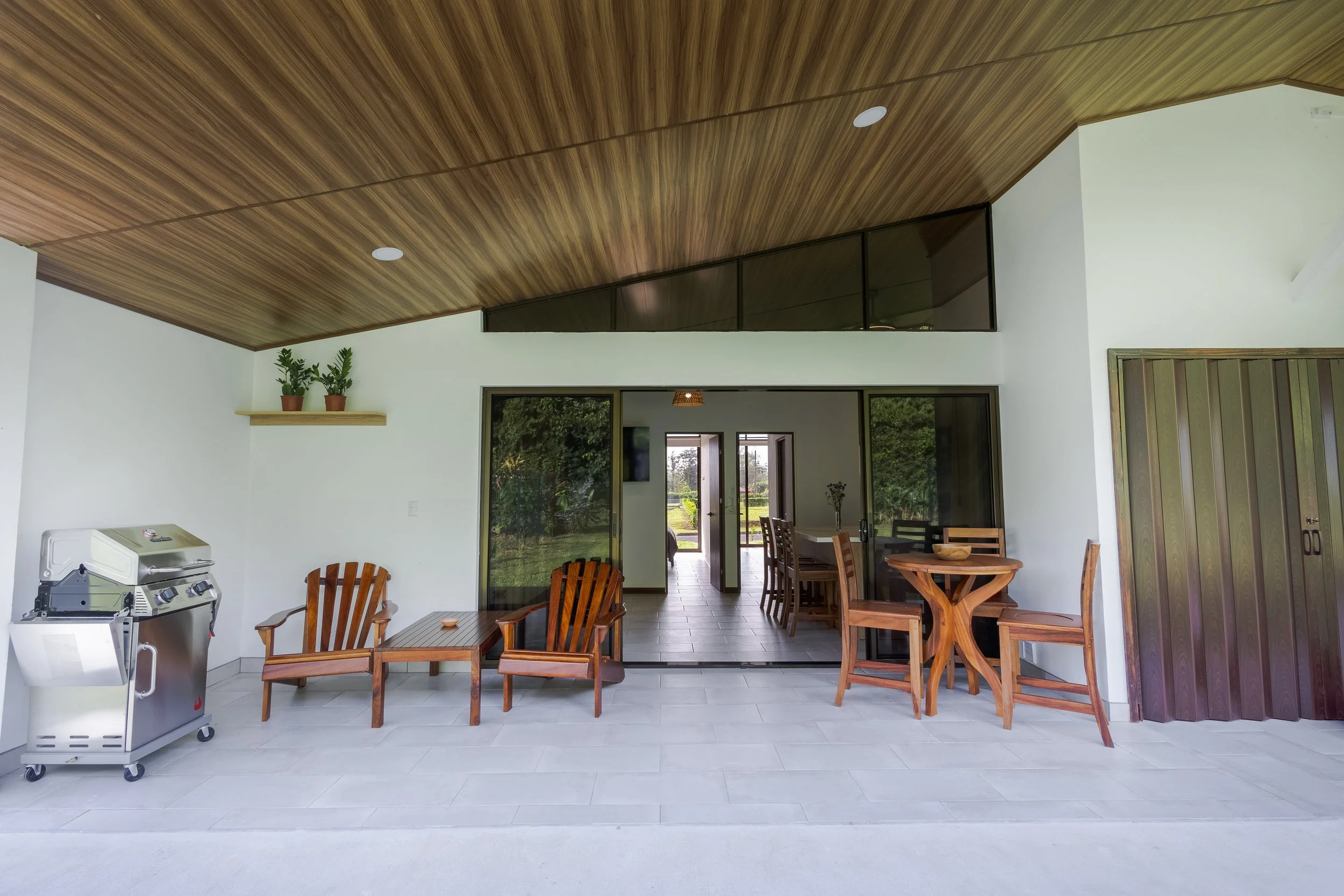 This image shows the interior of a modern house with white walls and a wooden ceiling. There are wooden chairs and a round table in the dining area, a sliding glass door leading outside, a small shelf with potted plants, and a barbecue grill on a whi