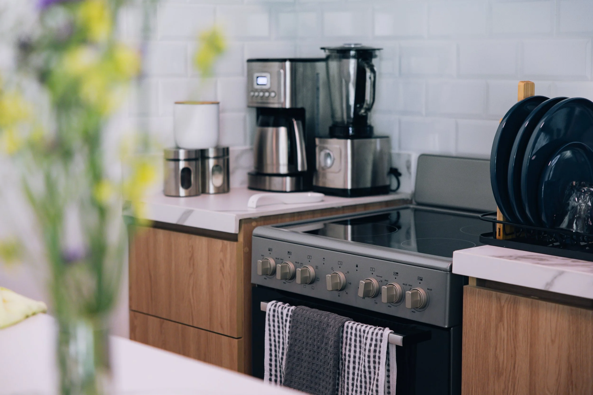 Kitchen with a white stove, wooden cabinets, and a gray and white striped towel hanging on the oven handle. There are small kitchen appliances on the countertop including a blender, coffee maker, and toaster. A partial view of a vase with green and p