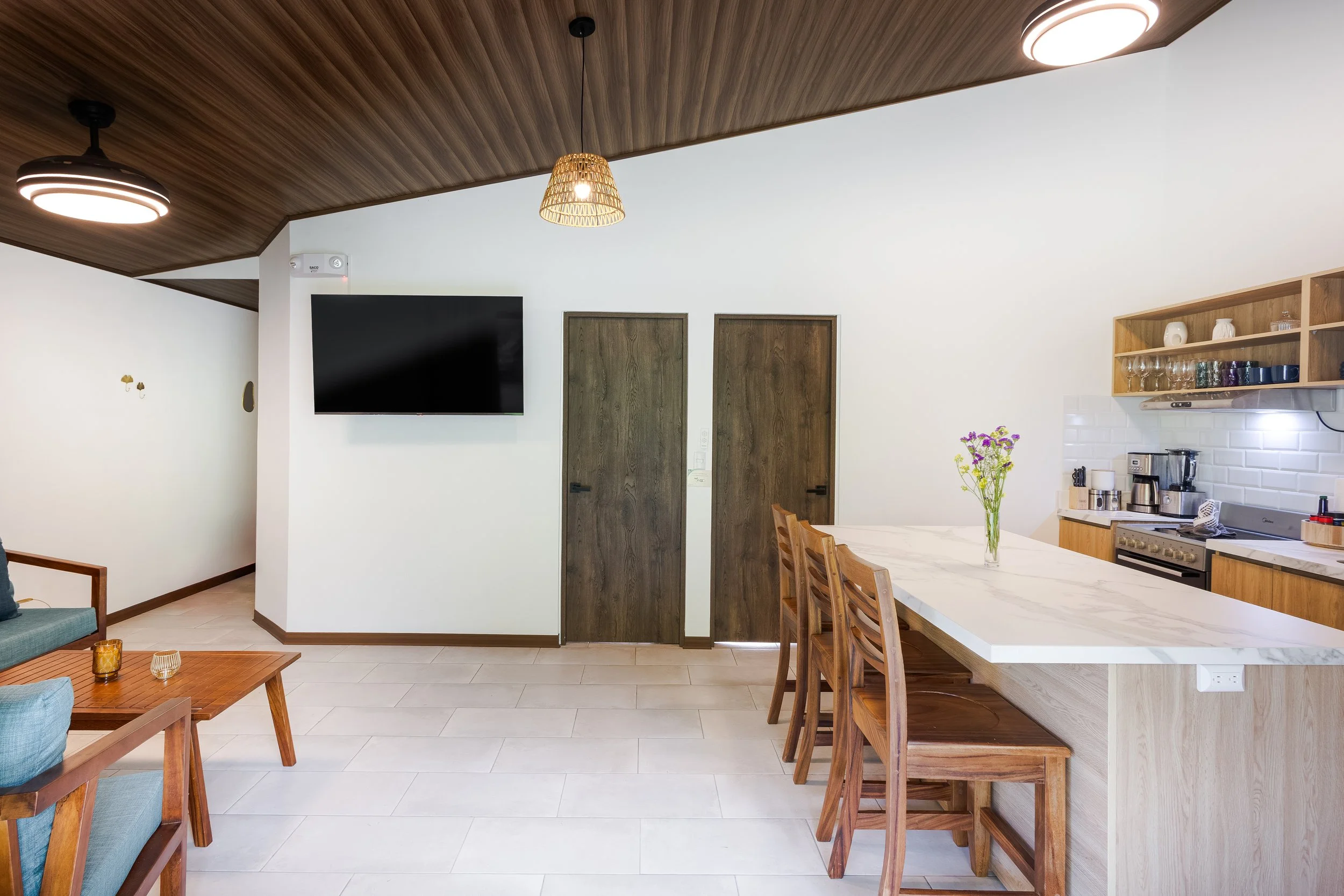 A modern kitchen and dining area with a wooden ceiling, white walls, a flat-screen TV, and a marble dining table with wooden chairs. There are wooden doors, open shelves with glassware, and a vase with flowers on the counter.