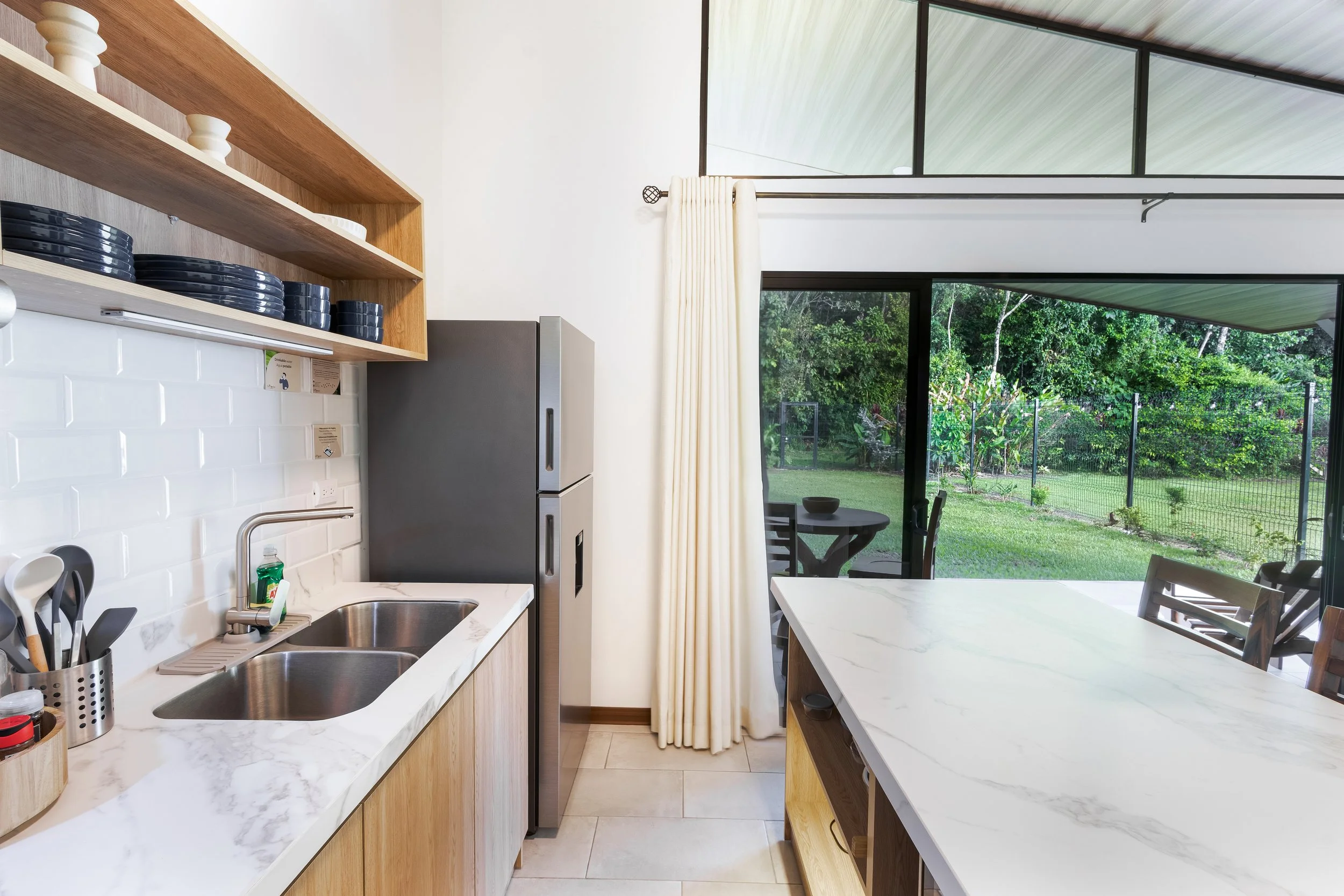 Modern kitchen with white countertops, wooden cabinets, stainless steel refrigerator, open shelf with dishes, and large glass sliding door leading to an outdoor yard with trees.