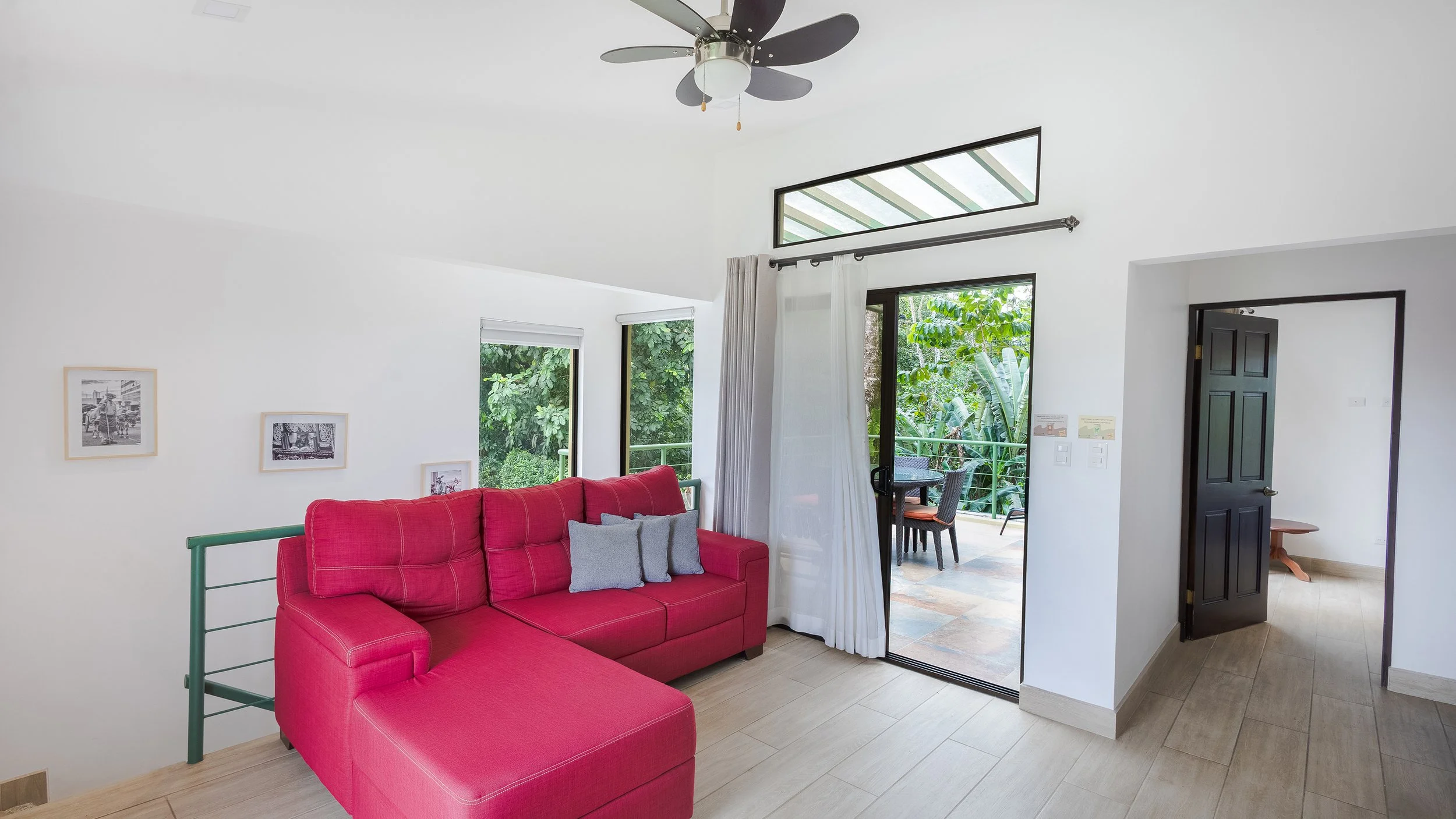 Living room with a red sofa, black door, large windows, and a sliding glass door leading to a balcony with outdoor furniture, surrounded by green trees.