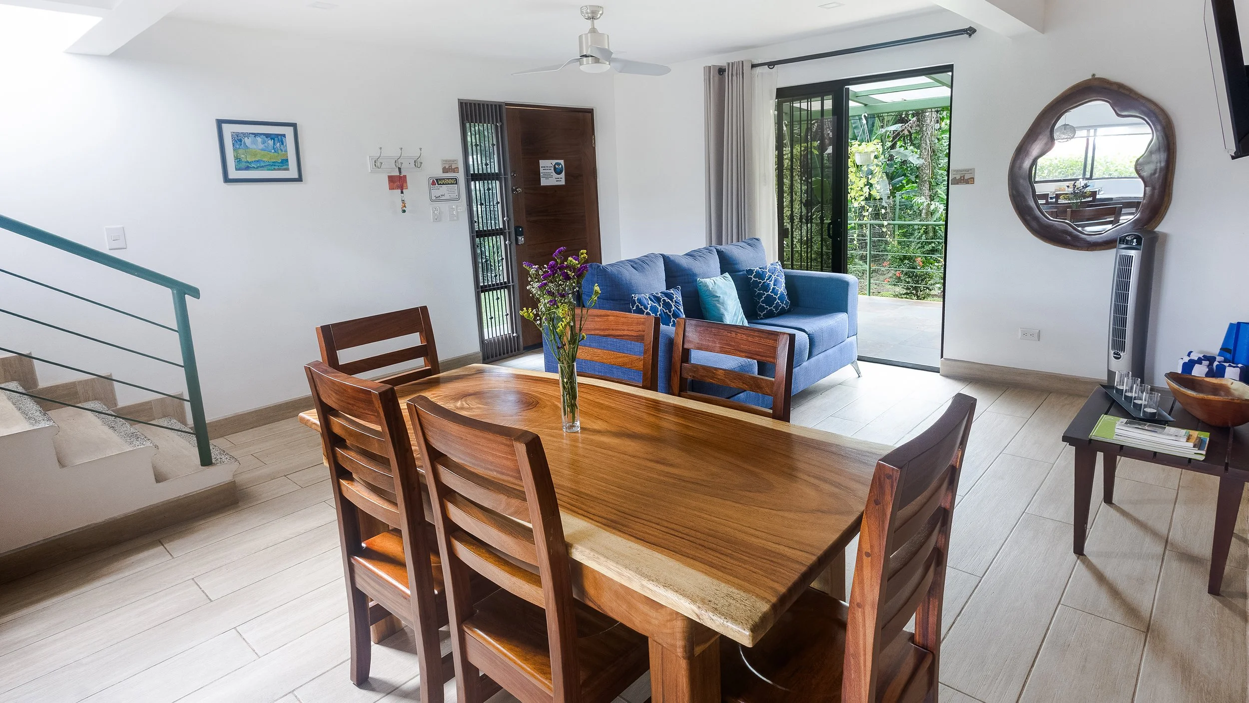 Living room with a wooden dining table and six chairs, a blue sofa with patterned pillows, sliding glass door with curtains, a mirror on the wall, a ceiling fan, and a view of greenery outside.