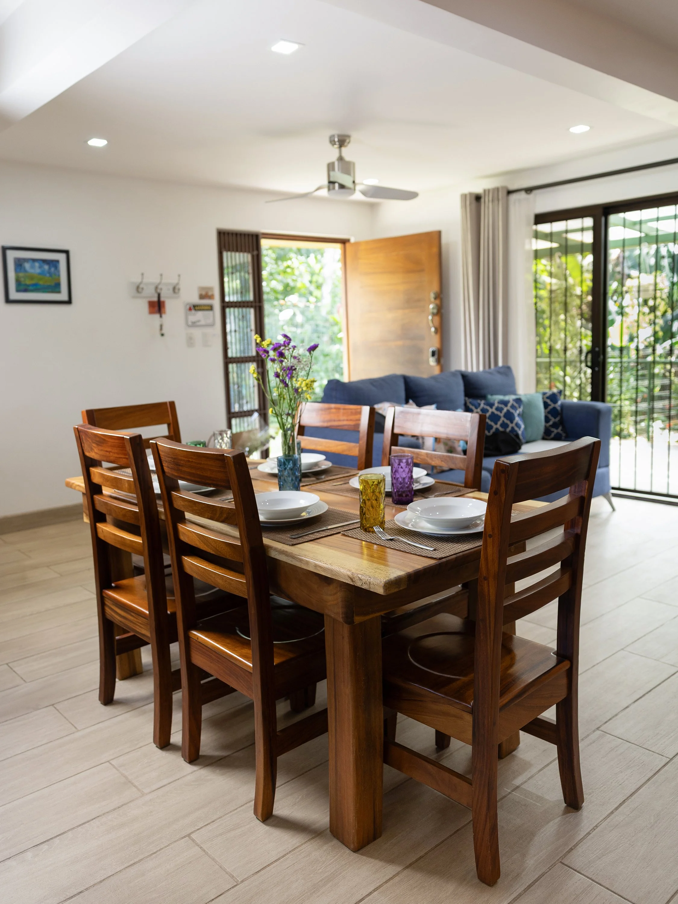 A dining area with a wooden table set for five, featuring white plates, colorful glasses, a vase with purple and yellow flowers, in a room with a light-colored floor, a blue sofa, a ceiling fan, and large sliding glass doors with curtains leading to 