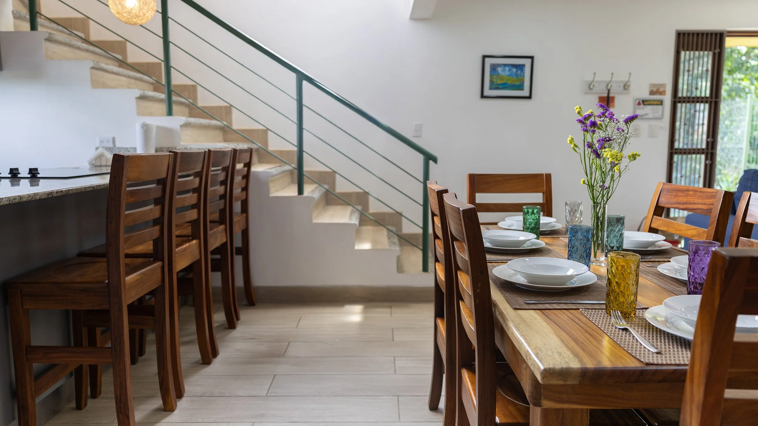 Dining room with a wooden table set with white plates, colorful glasses, and a vase with purple and yellow flowers. There's a staircase on the left with wooden stairs and green railing, and a kitchen counter with wooden chairs. The background include