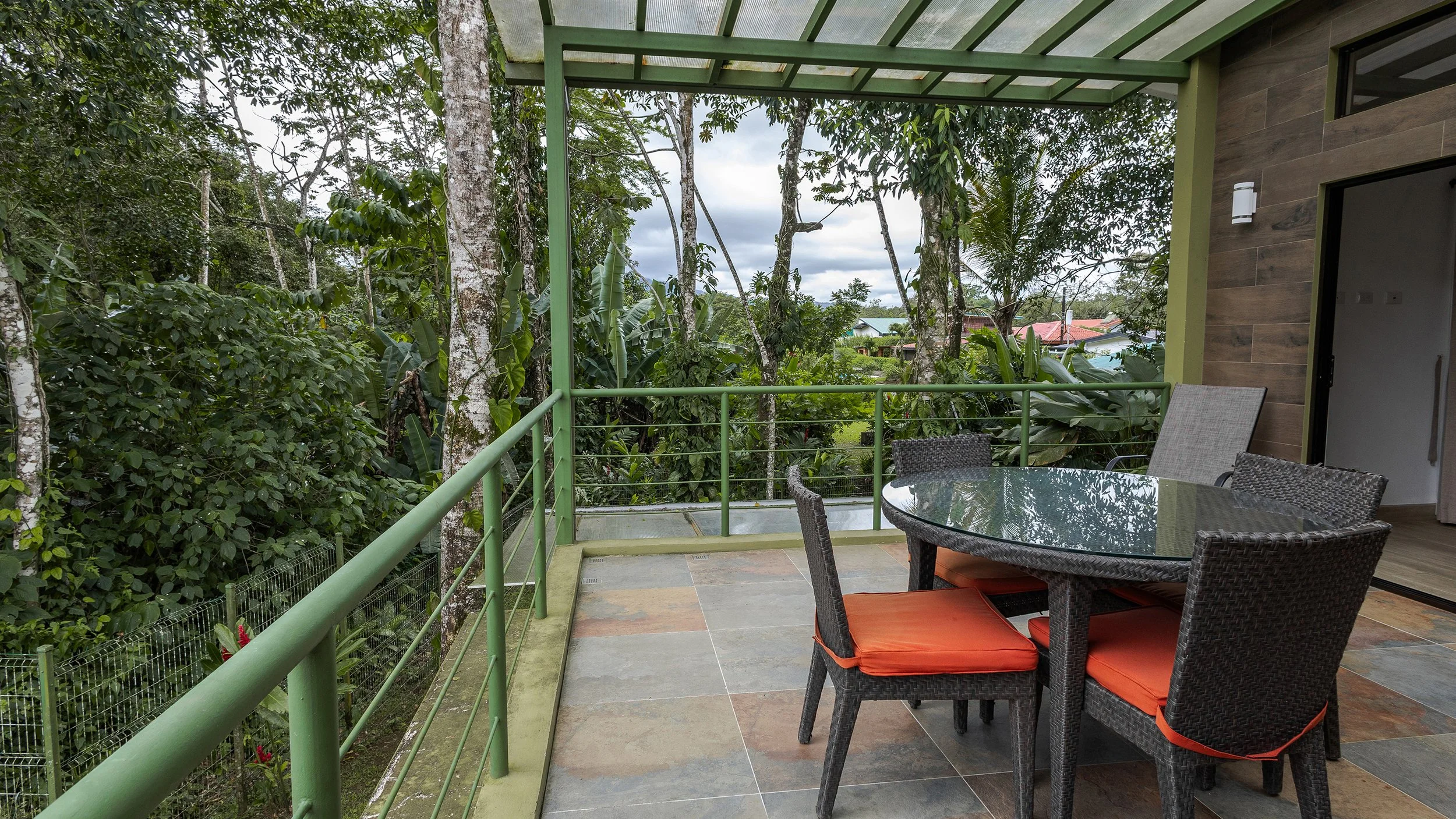 Balcony with glass-topped table and four wicker chairs with orange cushions, overlooking a lush green garden with trees and neighboring houses in the background.