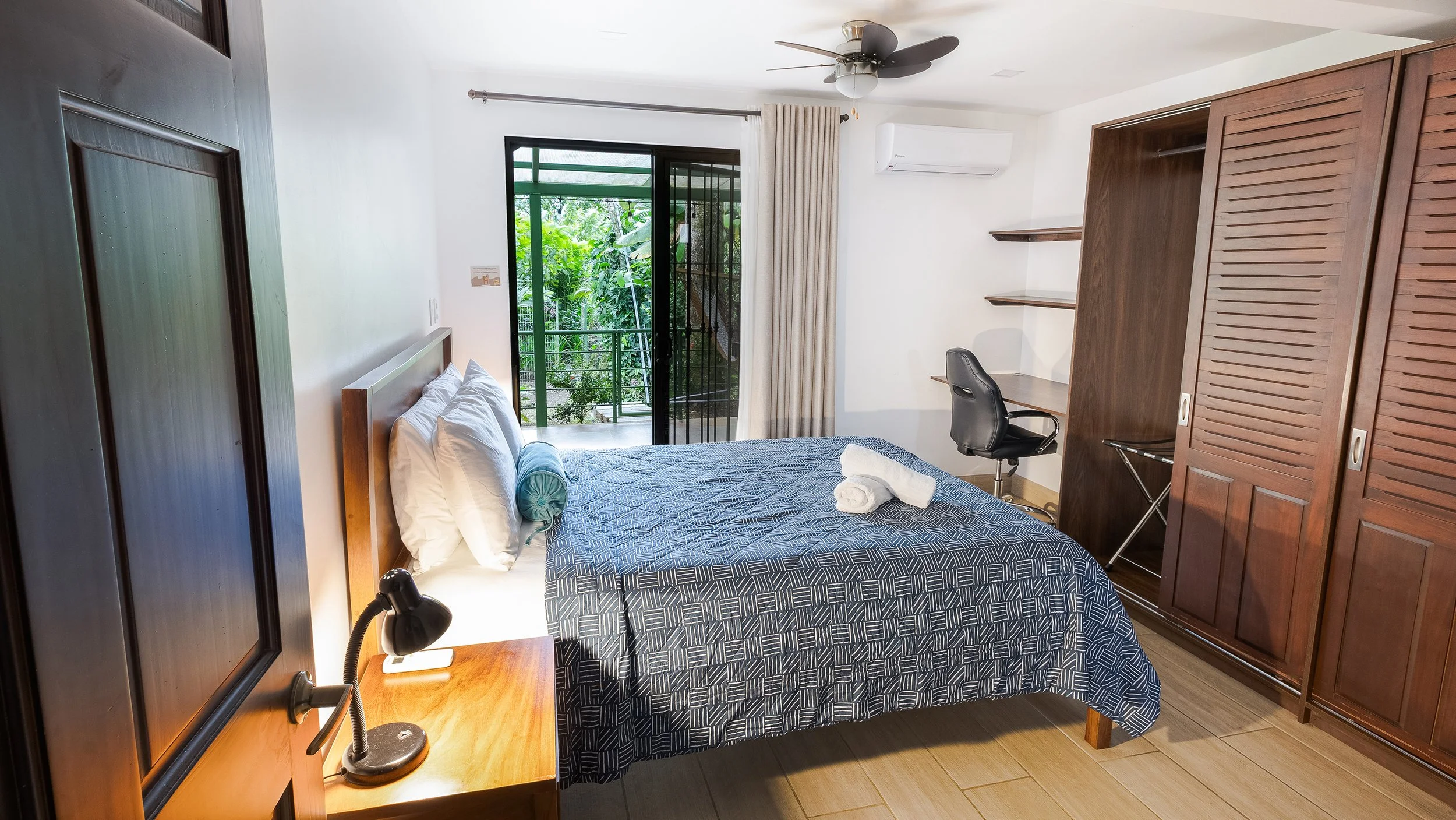 A bedroom with a sliding glass door leading to a balcony with greenery outside. The room has a bed with a blue patterned bedspread, white pillows, and two rolled towels on top. There is a wooden nightstand with a black lamp, a desk with shelves, a bl