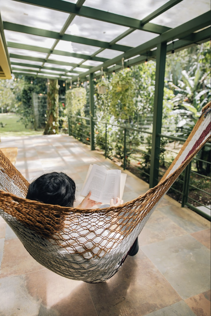 Person lounging in a hammock on a covered porch, reading a book surrounded by greenery and hanging plants.