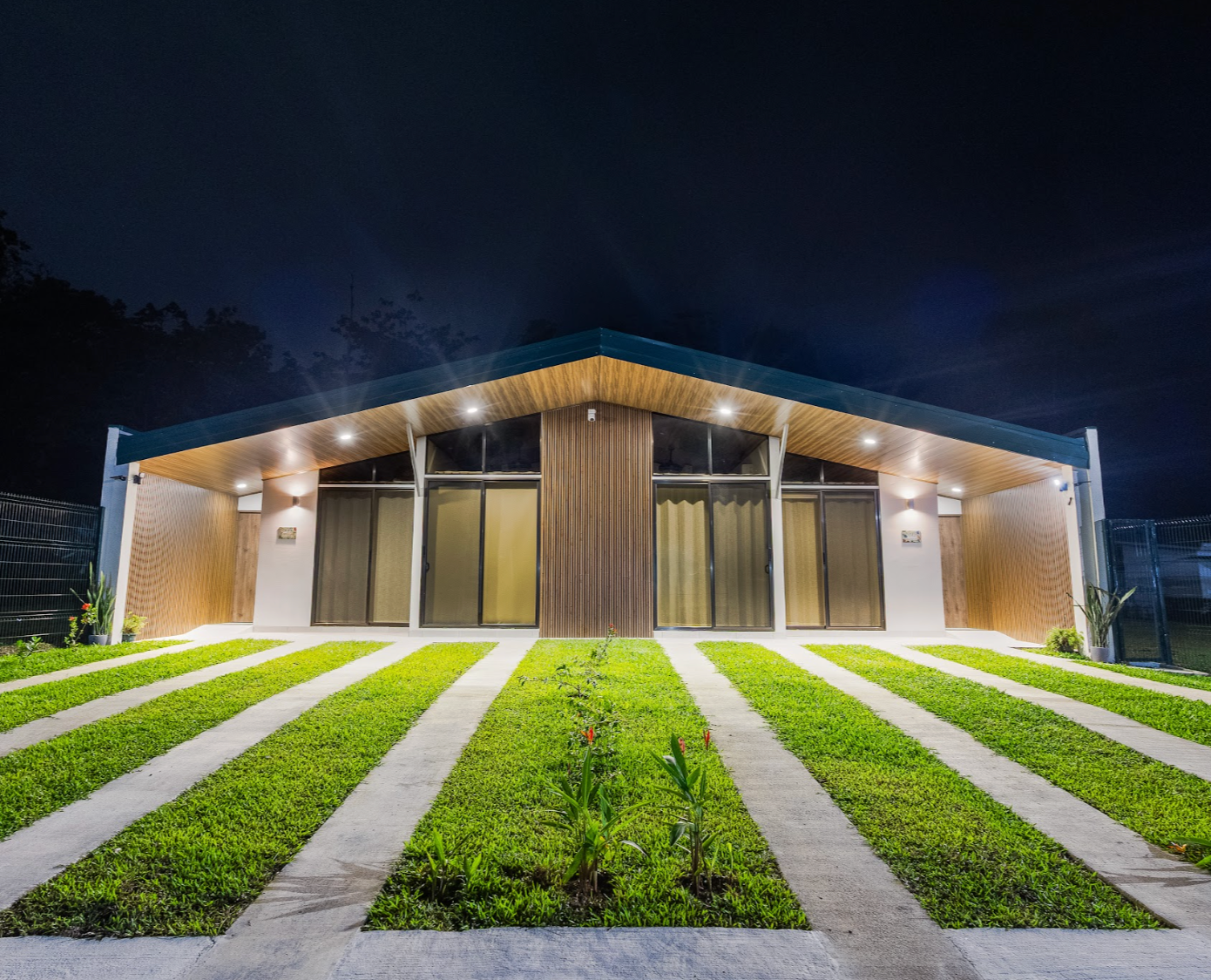 Modern house at night with illuminated outdoor lights, large glass windows, a sloped roof, and a front yard with green grass and concrete pathways.