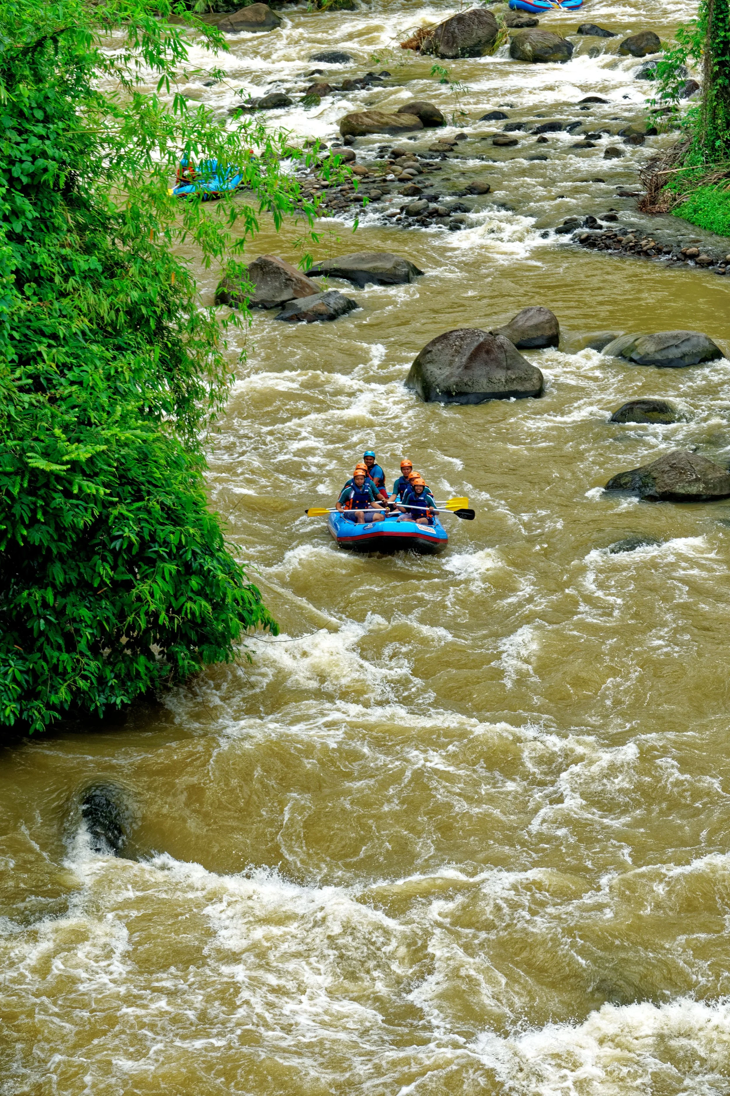 A group of people white water rafting on a river with brown water, surrounded by green foliage and rocks.
