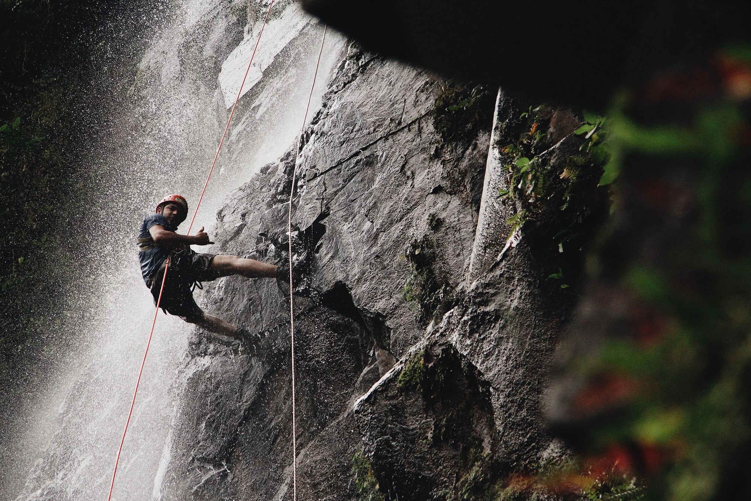 Person rock climbing on a wet, rocky surface with water spraying around, wearing a helmet and harness.
