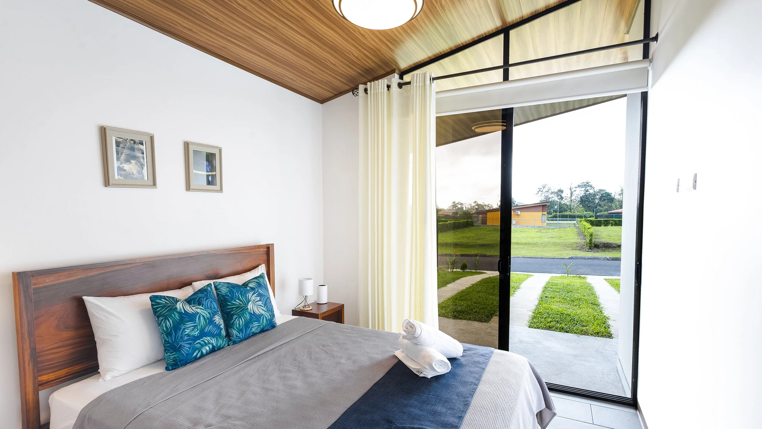 Bedroom with wooden headboard, blue tropical-patterned pillows, sliding glass door open to a grassy yard, white curtains, two bedside lamps.