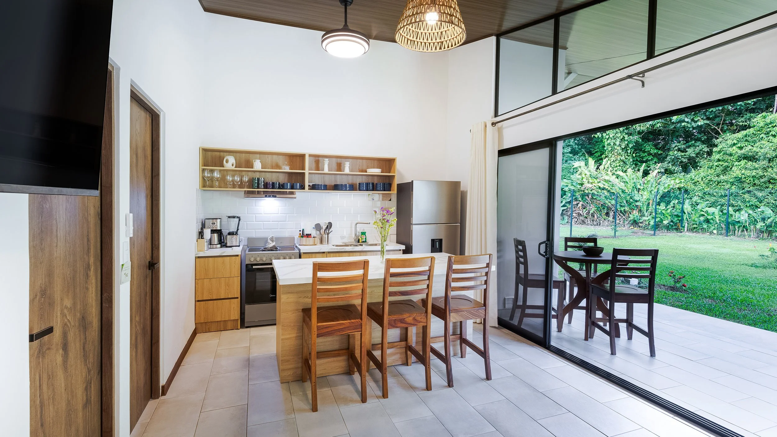 Modern kitchen with white walls, wooden cabinets, and a dining area with four wooden chairs and a table, adjacent to a sliding glass door opening to a lush green garden.